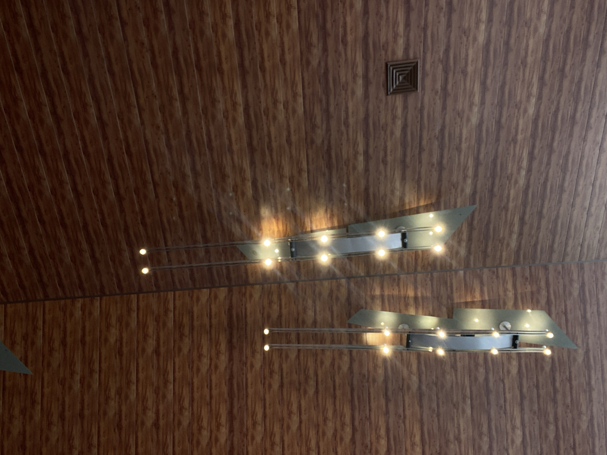 A low-angle shot shows a ceiling with two modern light fixtures and a square vent. The ceiling appears to be paneled with a wood-like material arranged vertically. The light fixtures are made of metal with a sharp, angular design and have several small light bulbs. The location is not evident in the image.