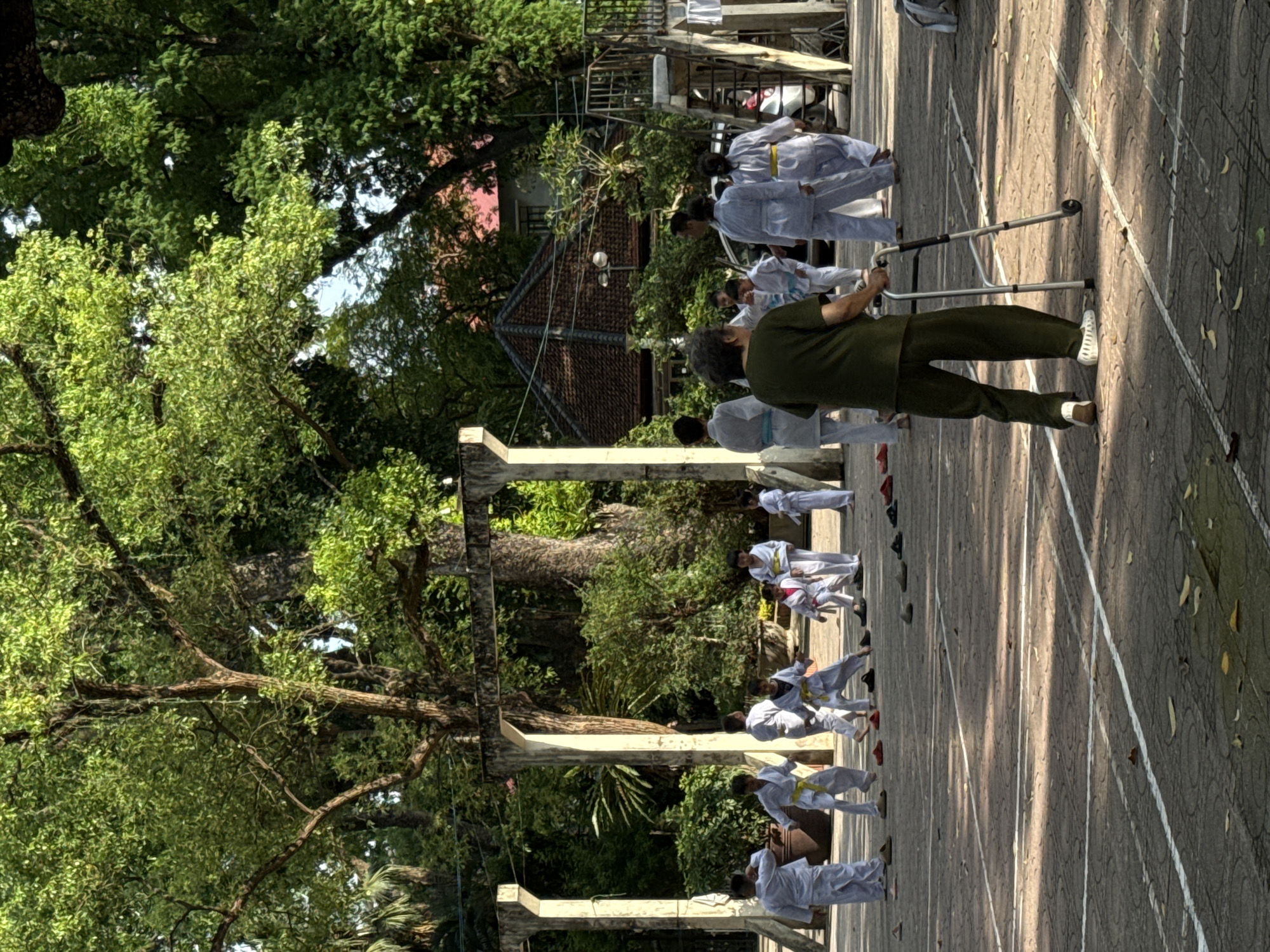 A horizontal outdoor shot shows a group of people practicing martial arts in Hà Nội, Vietnam.
The people are lined up in rows on a concrete surface marked with white lines, resembling a sports court. They are dressed in white uniforms, some with colored belts indicating their rank. Most of them are facing away from the camera, in various martial arts stances, while an older man in an olive drab uniform uses a walker and faces the camera, and appears to be instructing them.
The environment includes trees and architectural structures, possibly part of a park or public space. The lush greenery of the trees contrasts with the gray concrete, with light passing through the leaves. In the background, buildings with tiled roofs suggest the urban setting of Hà Nội. The scene is bathed in sunlight, casting shadows and creating a sense of depth.
