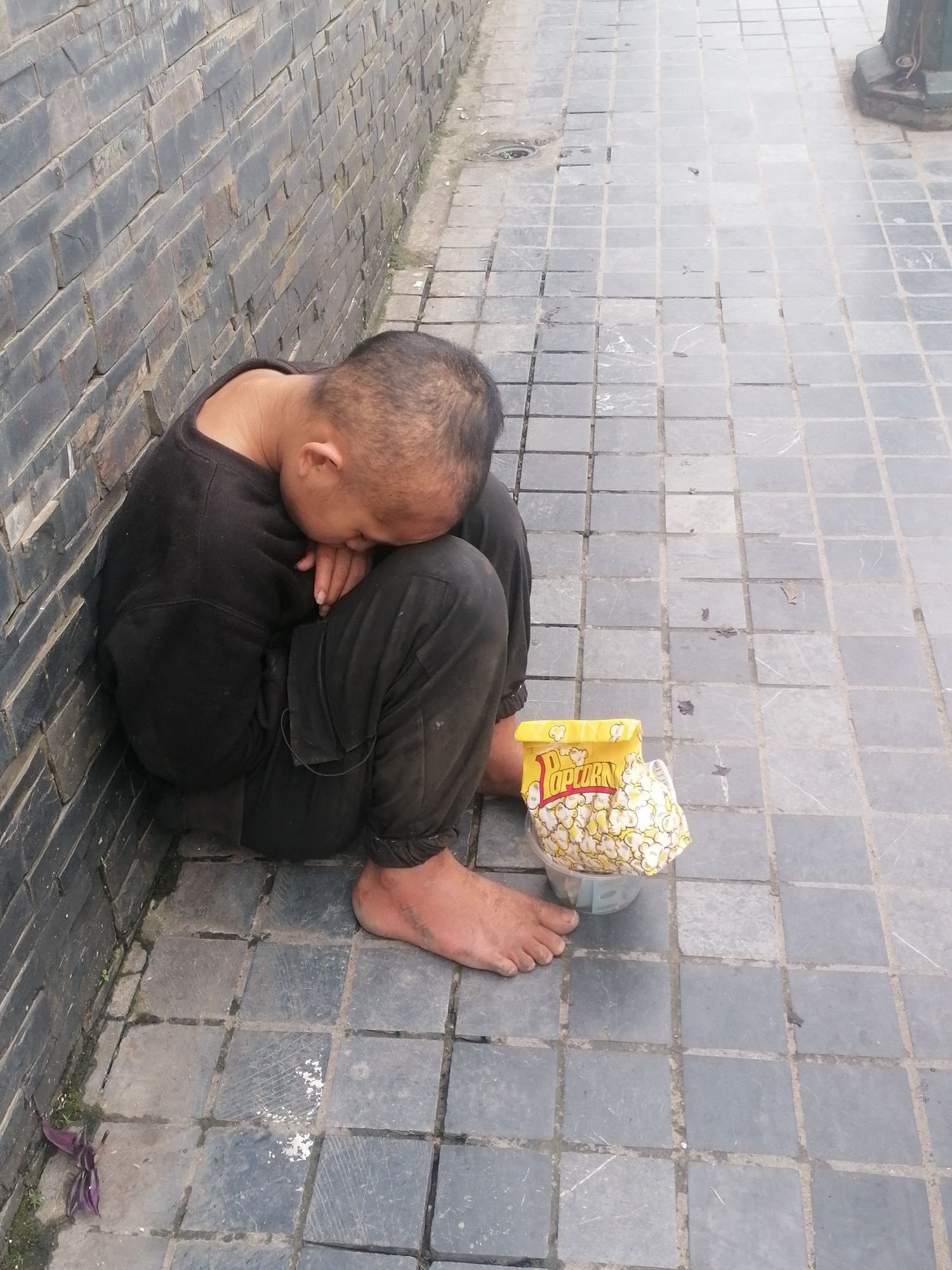 An individual is depicted resting or sleeping on the ground in what appears to be an outdoor urban or semi-urban setting in Sa Pa Ward, Vietnam. The person, who appears to be an adult male, is curled up with his head resting on his knees, leaning against a dark brick or stone wall. He is dressed in dark, possibly soiled, long-sleeved clothing and pants, and his feet are bare.
On the grey square-tiled ground beside him, a plastic container filled with popcorn is visible, partially covered by a yellow bag prominently displaying the word "Popcorn" in red text. The ground surface is paved with square tiles, showing some signs of wear and dirt, with small weeds growing along the base of the wall. The lighting suggests it is daytime, possibly overcast, as there are no distinct shadows. The scene conveys a sense of weariness and vulnerability, characteristic of someone in a difficult situation, possibly homeless.