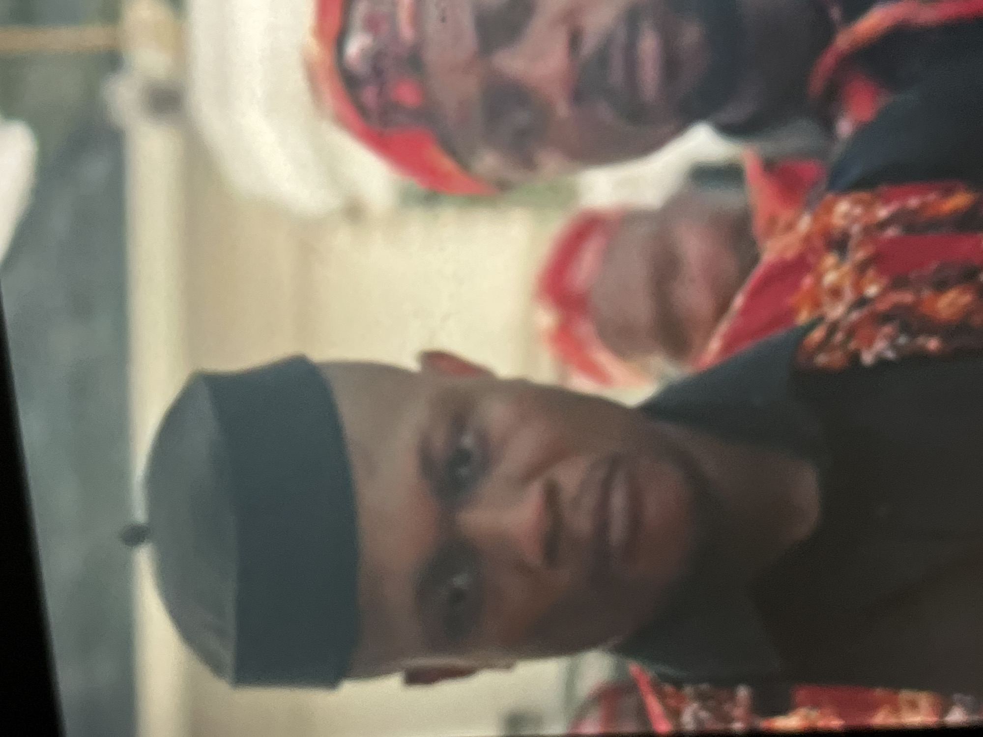 The image, taken in Abuja, Nigeria, captures three men in what appears to be a cultural or formal gathering. The photograph is vertically oriented, showing the subjects tilted. In the foreground, a man with a shaved or very short haircut is sharply in focus. He wears a dark, possibly black, cap (resembling a Kufi) and a dark-colored garment. He looks directly at the camera with a neutral to slightly serious expression.
Behind him, positioned to his right and slightly out of focus, are two other men. They are adorned in vibrant traditional attire, featuring head coverings or caps made of patterned red and orange fabric, and garments with intricate designs in similar hues. Their expressions are less discernible due to the blur. The background is soft and indistinct, suggesting an indoor setting or a shaded outdoor area, characterized by light-colored, blurred structures or walls. The distinct traditional clothing strongly suggests a cultural event or a traditional gathering in a West African context. There is no visible text in the image.