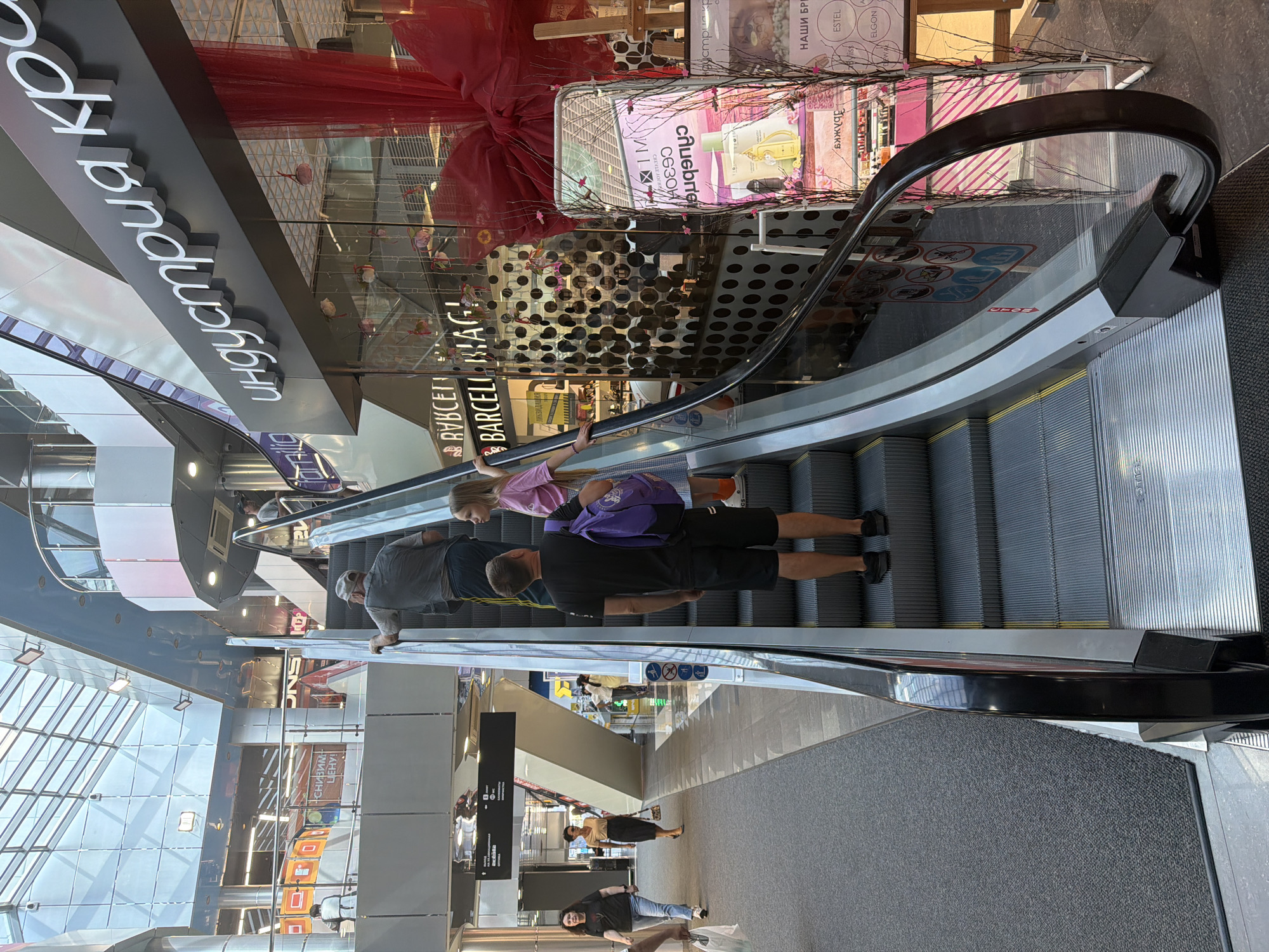 The image shows an indoor scene, likely a shopping mall, with multiple levels visible. An escalator is a prominent object, with several people on it. On the escalator, a young girl in a pink shirt and a purple backpack is riding with an adult man in a black shirt and shorts. Another adult man in a grey shirt and cap is also on the escalator. In the background, there are shops and signs with text in Russian, including "индустрия кр", "ВУВСЕГ", "BARCELI BAG!", and "ЛИКВИДАЦИЯ". Another sign reads "снизим LEHY!" and "UNS 10 STIS". Further in the distance, more people are visible on different levels of the mall. The setting appears to be well-lit, suggesting it's daytime, and there are no indications of the weather. The overall atmosphere seems casual and everyday, with people going about their activities in a commercial space.