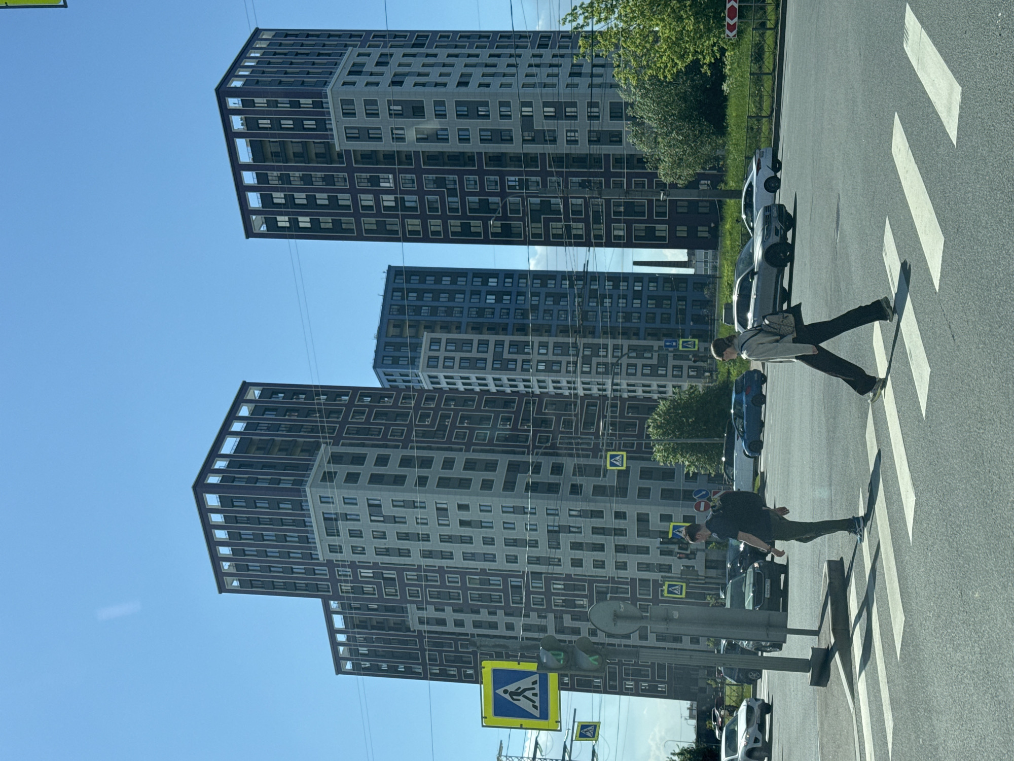 The image displays a bright, sunny daytime street scene in an urban area, identified as Sinyavino, Russia. Two pedestrians are actively crossing a zebra crossing on an asphalt road. The person in the foreground wears a light-colored jacket and dark trousers, carrying a shoulder bag, while the second person, slightly behind, is dressed in dark clothing. Both cast distinct shadows on the road, indicating strong sunlight.
Several vehicles are visible on the road, including a light-colored SUV and a dark sedan. To the right of the road, green foliage and a dark metal fence line the street.
In the background, multiple tall, modern apartment buildings dominate the skyline. These structures feature a contemporary design with a pattern of alternating dark and light panels across their facades. The sky is clear and blue.
Various road signs are present, including several yellow diamond-shaped pedestrian crossing signs with a black figure, and a red triangular sign with a white background and a black chevron pointing right, indicating a priority road. A traffic light with a green pedestrian signal is also visible, along with overhead utility wires stretching across the frame. The scene portrays a typical urban environment with ongoing pedestrian and vehicle traffic amidst modern architecture.