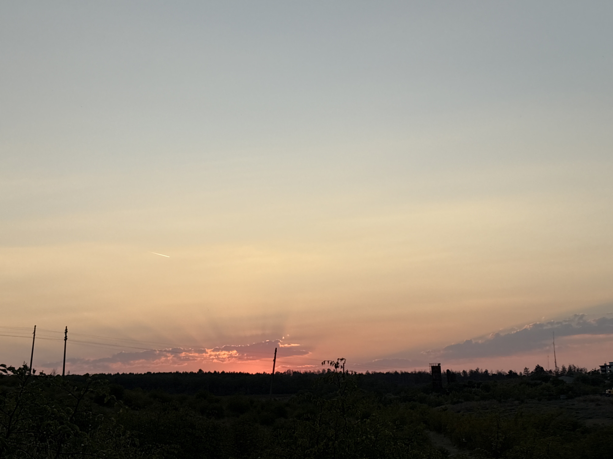 This is a photograph of a rural landscape at sunset in the city of Tankovo, Bulgaria. The sky is a gradient of soft colors, transitioning from a pale blue at the top to hues of orange and pink near the horizon. Rays of sunlight emanate from behind a line of trees and clouds, creating a dramatic effect. In the foreground, silhouetted trees and vegetation form a dark, dense line across the bottom of the frame. Two utility poles with wires are visible in the middle ground, and further in the distance, there are hints of structures and trees, including a dark tower-like object on the right side of the image. A contrail from an aircraft is visible in the upper portion of the sky.