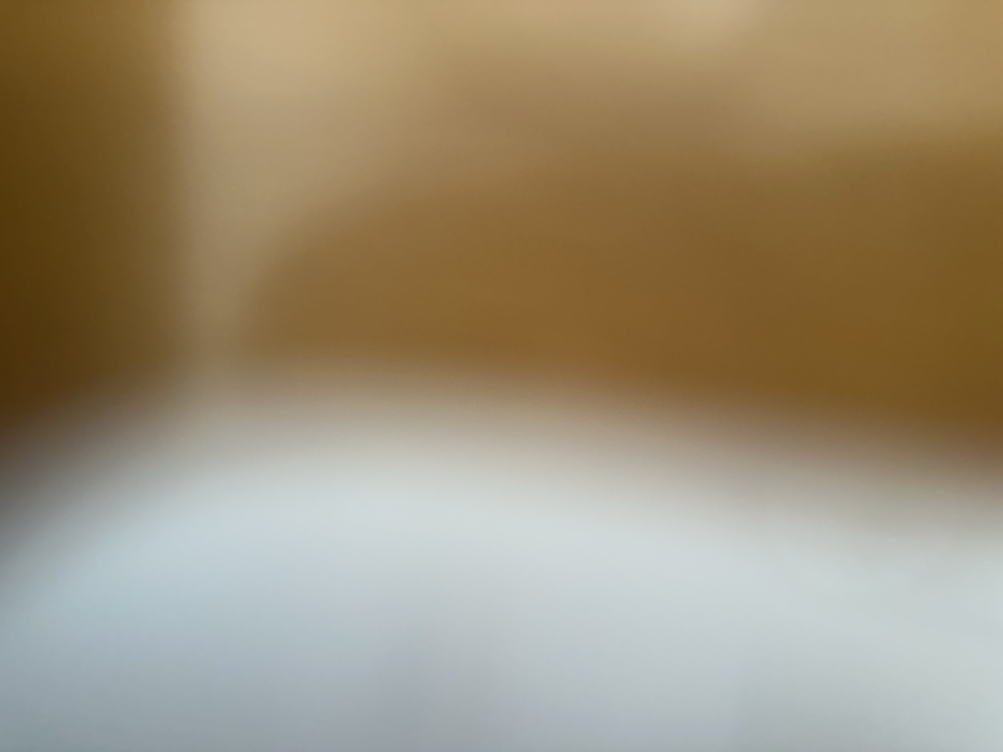 The image is an abstract, out-of-focus depiction of a surface with subtle color gradients. The upper portion of the image is dominated by warm, golden-brown tones, suggesting a material like wood or a textured wall. This area is illuminated unevenly, with a brighter, lighter patch on the left, indicating a light source or a change in surface reflectivity. The lower portion transitions into lighter, cooler, grayish-white tones, creating a stark contrast with the upper section. This area is soft and diffused, possibly representing a different material or an area in shadow. There are no discernible subjects, objects, or activities taking place. The overall impression is one of texture and light rather than a concrete scene. The image lacks any specific details related to time of day, weather, or location cues. No text is visible. The provided crops offer a closer, yet still blurred, view of these color and light variations.