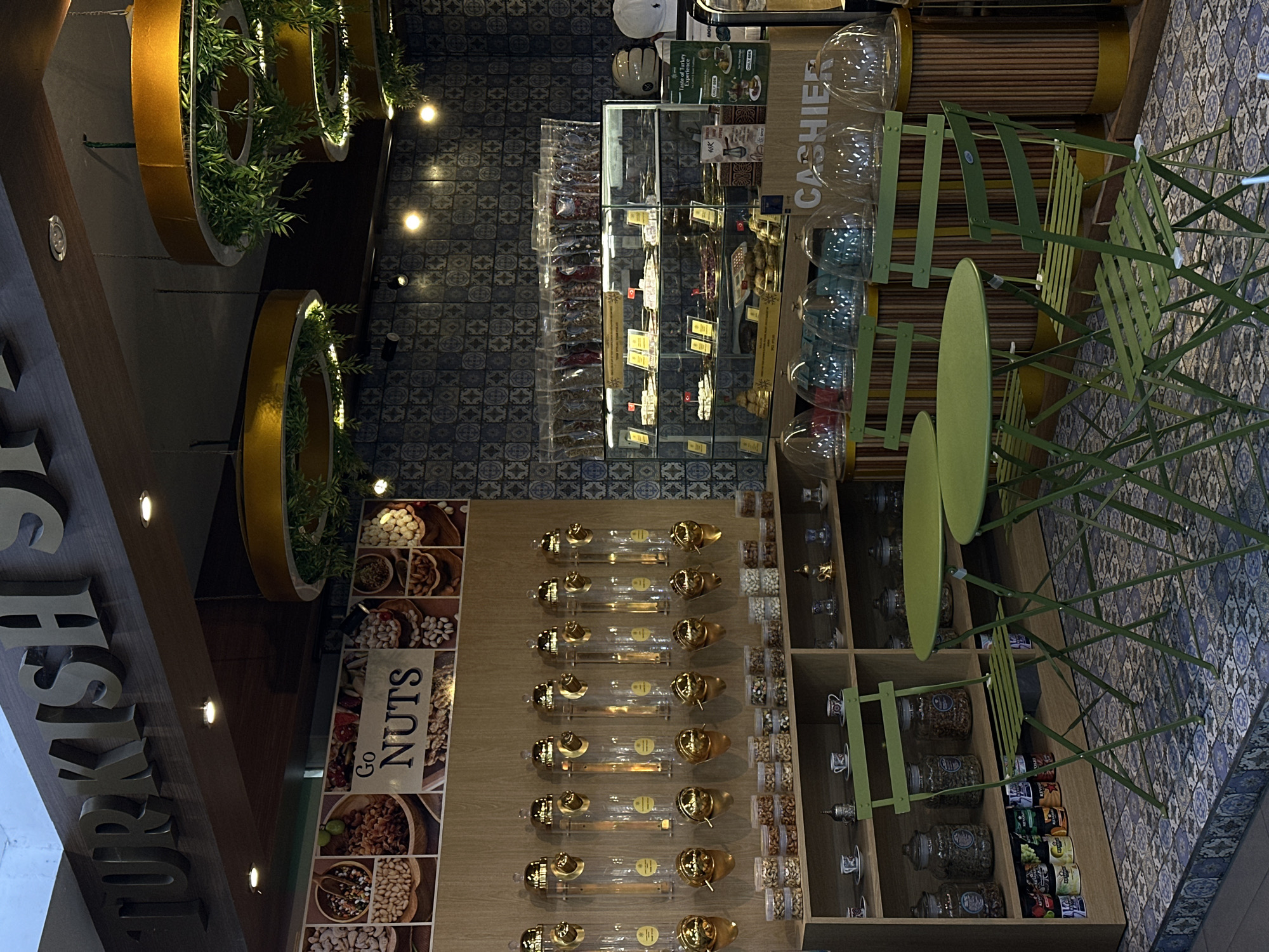 The image depicts an interior shot of a shop, likely a Turkish nut store, in an unknown area of North Kuta, Indonesia. The shop features rows of clear tubes filled with nuts, a glass display case containing various products, and a "Cashier" sign accompanied by large, clear storage containers. Green metal chairs and a round table are positioned near the cashier area. The decor includes decorative rings with greenery hanging from the ceiling and a sign with the words "Go Nuts." The walls are tiled with a blue-and-white patterned design.