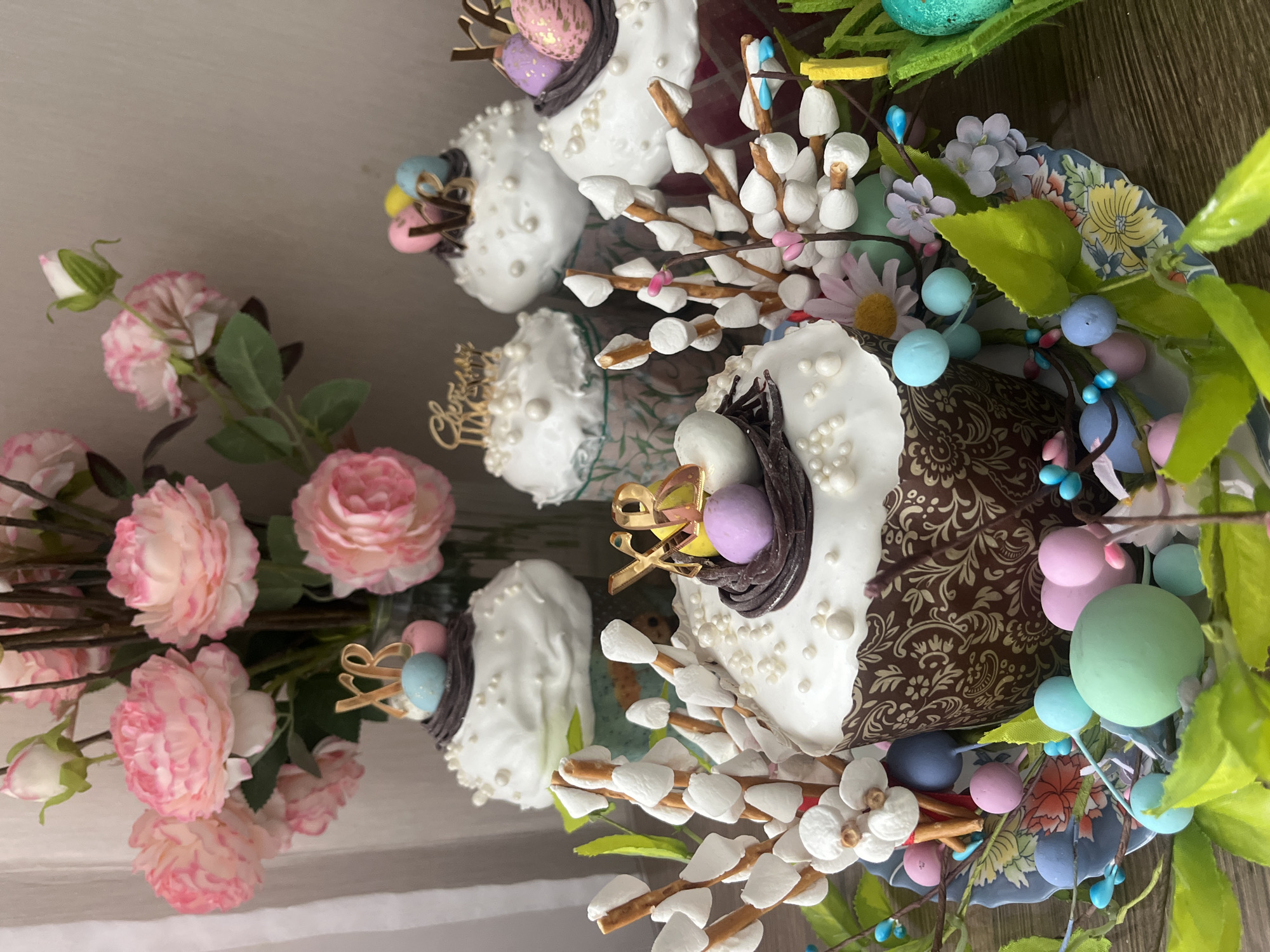 The image features a collection of Easter cakes, decorated with white icing, pearl-like sprinkles, and small nests containing colored eggs. Some cakes have additional decorations like golden "XB" (Christ is Risen) symbols. Willow branches with marshmallow-like buds are arranged around the cakes, along with other artificial flowers and greenery. To the left, there are pink roses. The objects are placed on a surface that appears to be wooden. There is no information to suggest the location is Nizhnevartovsk, Russia.
