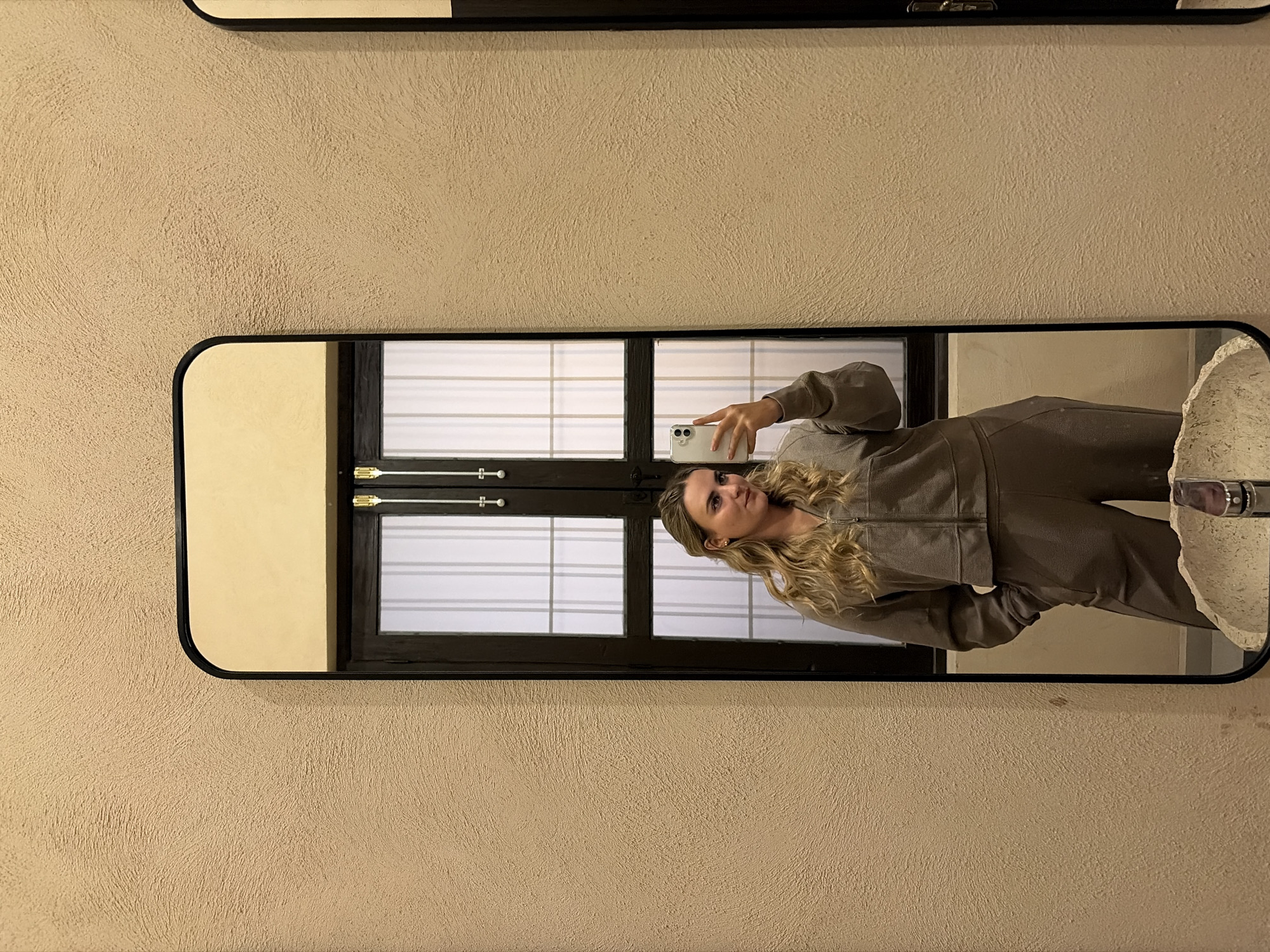 A medium shot depicts a mirror with a black frame hanging on a beige textured wall. A person is reflected in the mirror, holding a smartphone and taking a selfie. The individual has long, wavy blonde hair and is wearing a brown, zip-up jumpsuit. The reflection also shows a portion of a window with horizontal blinds and a white sink. The scene appears to be indoors, likely in a bathroom or dressing area. The city location is not discernible from the image.