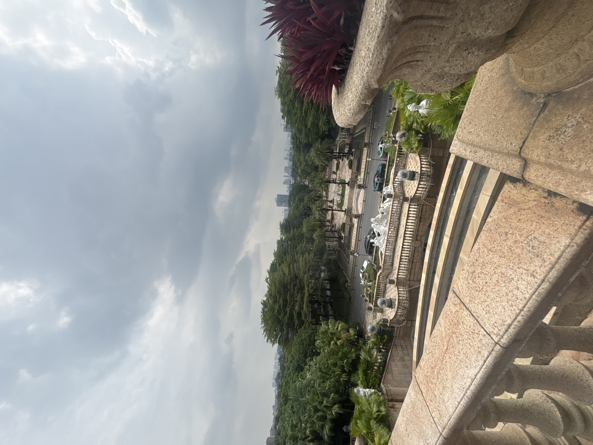 The image captures a view from an elevated vantage point, looking out over a city landscape in Nanhai District, China. In the foreground, a textured stone railing and balustrade suggest a terrace or balcony. Beyond this, a tiered stone staircase descends into a landscaped area.
The middle ground features a lush, green environment with numerous trees and palm trees. A road runs through this area, with two dark-colored cars and one lighter-colored car visible. Several white sculptures, some appearing to be human figures and others possibly animalistic, are scattered throughout the landscaping, some positioned near the road and others on platforms. A decorative fountain or water feature is also suggested.
In the background, a dense collection of trees forms a green expanse, leading to a city skyline in the distance. Tall buildings, some with cranes indicating ongoing construction, rise against a cloudy sky. The sky itself is overcast with a mix of light and dark gray clouds, suggesting a day with potentially intermittent rain or a generally diffused light. A potted plant with striking red foliage is prominently positioned in the far right of the frame, adding a splash of vibrant color to the scene.