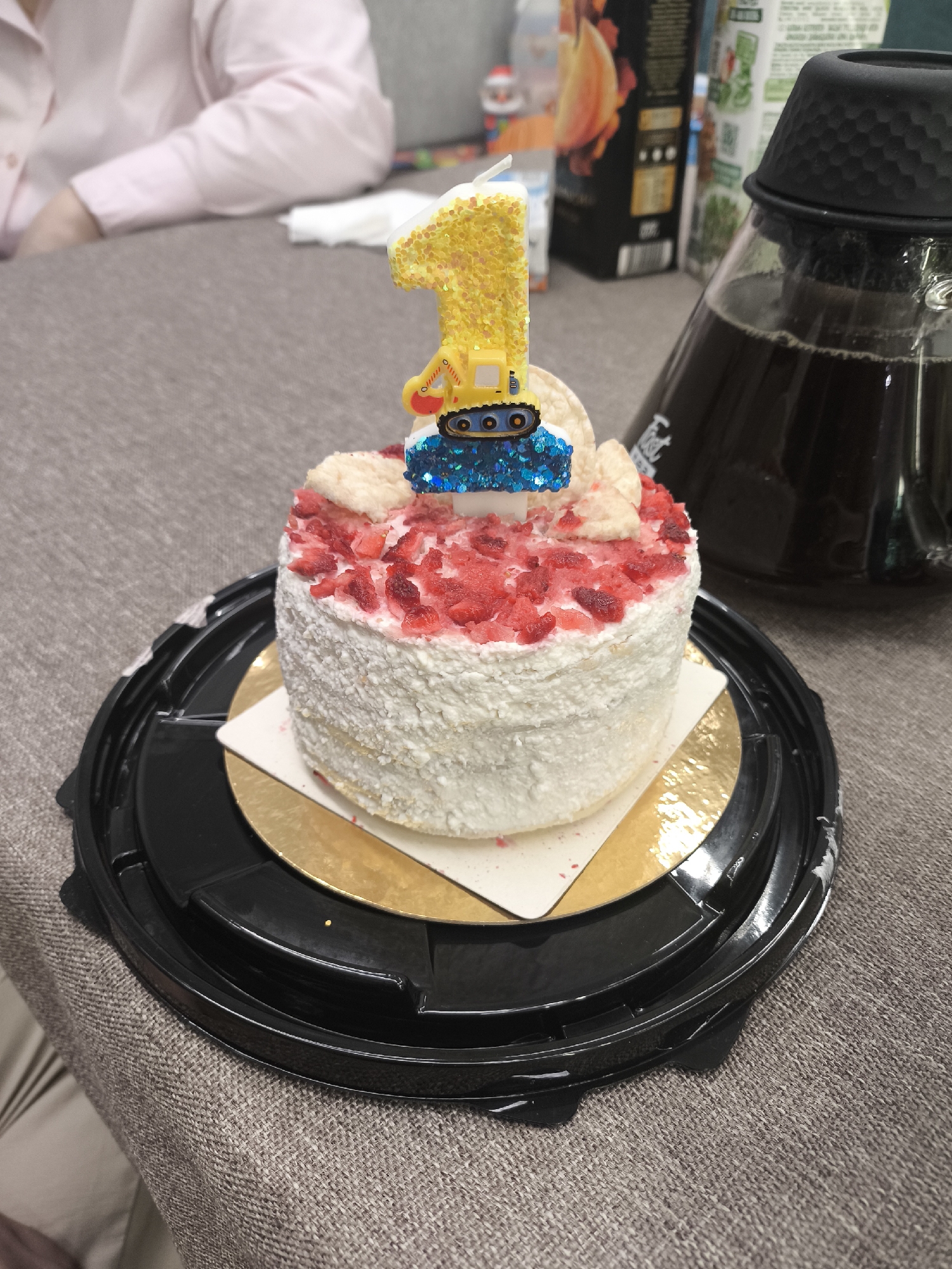 The image shows a small birthday cake on a black plastic plate placed on a textured beige surface, possibly a tablecloth. The cake is white, coated with what appears to be white crumbs and decorated with small, red pieces on top. On the top of the cake, there is a birthday candle in the shape of the number "1" with a small, yellow and blue construction vehicle figure. To the right of the cake is a large glass carafe containing a dark liquid. The background includes a person wearing a light pink shirt and blurred objects. The location appears to be indoors. There is no evidence to suggest the location is in Moscow, Russia.