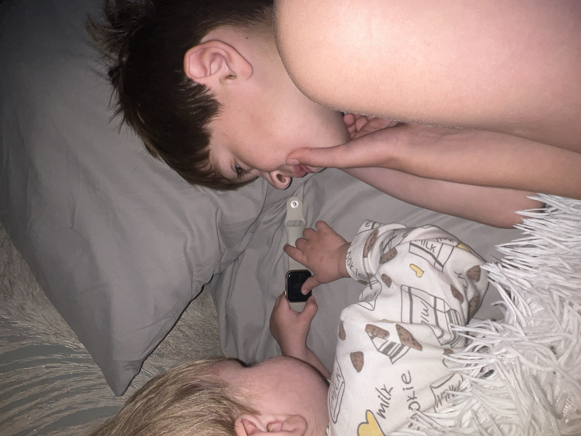 Two children are lying on a grey bed, one with brown hair and one with blond hair. The brown-haired child is on his side with his face near the other child's face. He is touching his chin. The blond-haired child is lying on his back with his hand holding an Apple Watch. The child is wearing a white pajama top with drawings of milk and cookies. The room appears to be dimly lit, and a white fringe is partially covering the child's body. The city of Noviy, Russia is not visually identifiable in the image.