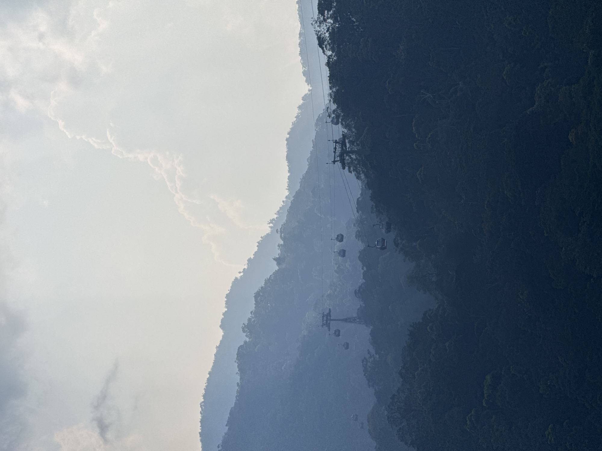 The image shows a view of the Bà Nà Hills cable car system in Đà Nẵng, Vietnam. The scene is dominated by layered mountain ranges, shrouded in varying degrees of mist and atmospheric perspective. The cable car system, with its towers and gondolas, stretches across the mountainous landscape. The foreground consists of dark, densely vegetated slopes, contrasting with the lighter, hazy background of clouds and sky. The orientation of the photograph is rotated, with the sky appearing on the left side of the frame and the ground on the right.
