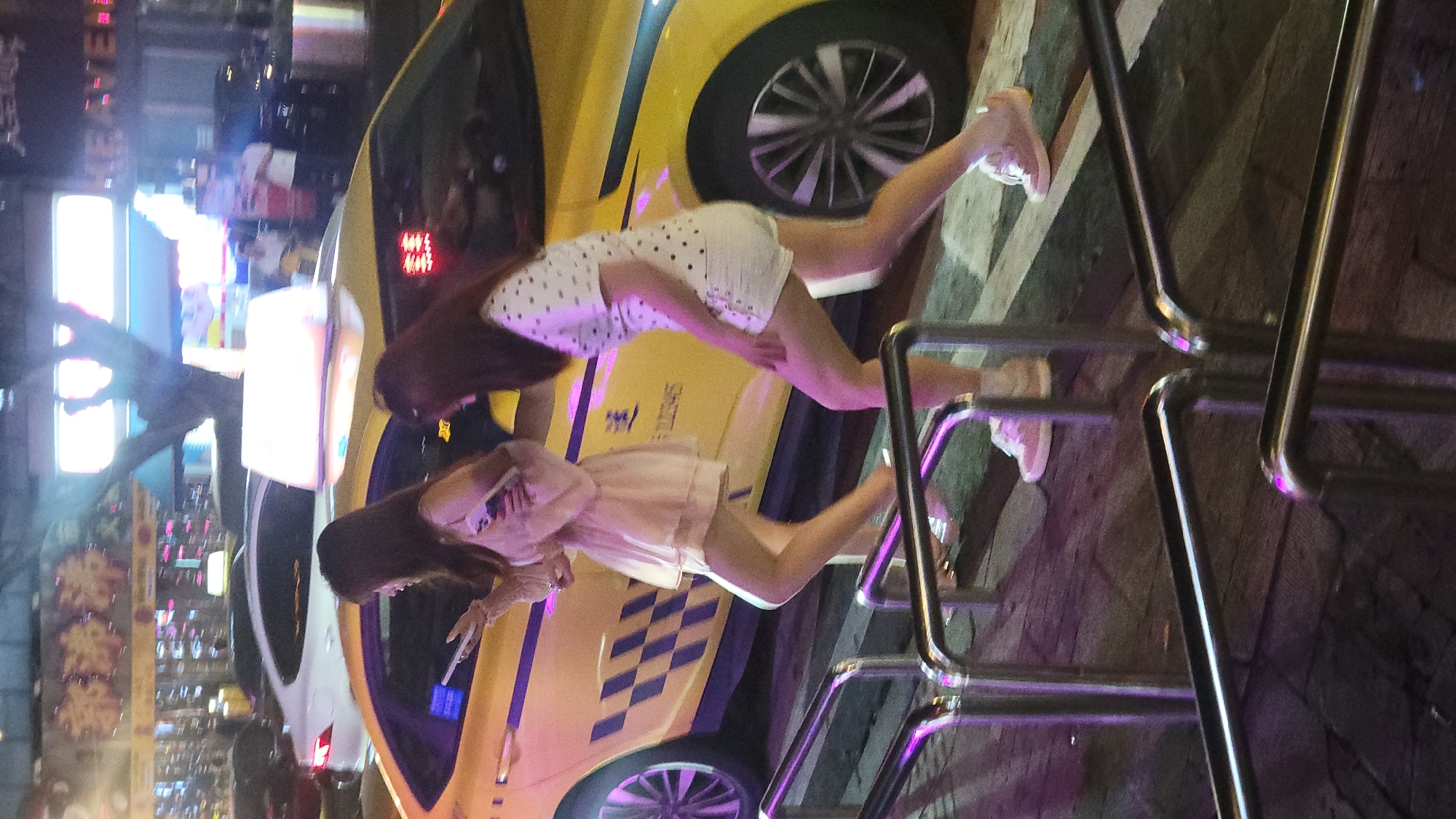 Here is a detailed description of the image:
**Scene:** An urban scene featuring two women posing in front of a yellow taxi. The photo appears to be taken at night, with city lights visible in the background.
**People:** Two young women are the main subjects. One woman is wearing a white polka dot dress. The other woman is wearing a light-colored dress. Both are wearing sneakers and are holding smartphones.
**Objects:**
* A yellow taxi cab with checkered detailing.
* Metal railing in the foreground.
* City lights and signage in the background, suggesting a commercial or downtown area.
**Actions and Interactions:** The two women are posing for a photo or video in front of the taxi. They appear to be interacting with their smartphones, likely taking pictures of themselves or each other.
**Environment:** The setting is an urban environment, potentially a street corner or a parking area. The background lights and signs suggest a lively, possibly touristy, area at night.