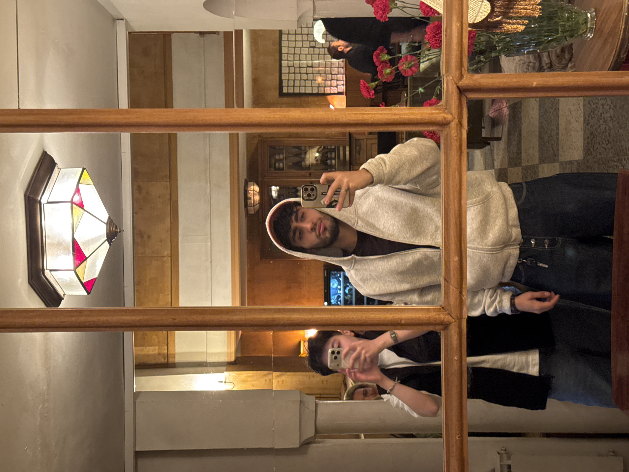 A young person wearing a gray hooded sweatshirt, dark jeans, and a dark shirt is holding a mobile phone, presumably taking a photograph of their reflection in a divided wooden-framed mirror. Another person with black hair, wearing a black and white top, is also holding a mobile phone and appearing to take a photo of the same mirror. In the background, there are flowers in a vase, decorative lighting, and portions of a striped wall, suggesting an interior space, possibly in Yerevan, Armenia. A stained glass lamp is visible in the reflection.