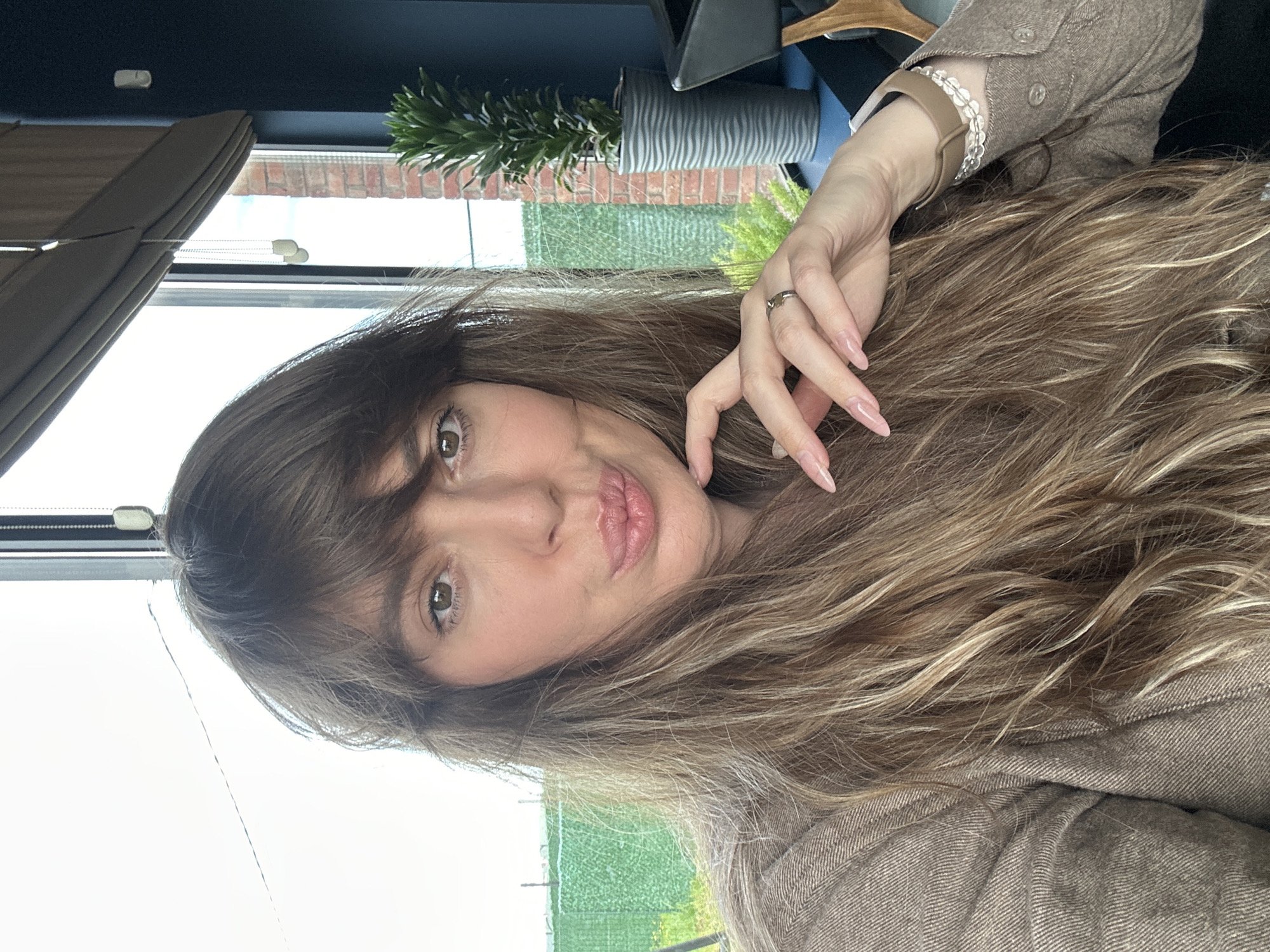 A woman with long, wavy brown hair is posing for a selfie. She is wearing a brown shirt and has a ring on her left hand. Behind her is a window with a view of a green field and a brick building. There is a plant in a gray pot on the windowsill. The location appears to be indoors, possibly a cafe or home in Marneuli, Georgia, based on the type of buildings and vegetation visible through the window.