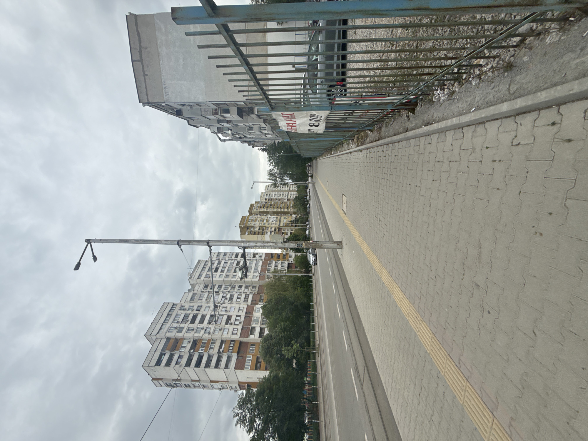 The image depicts a street scene in Sofia, Bulgaria, characterized by aging, Soviet-era apartment buildings lining a thoroughfare. The buildings are mostly white with orange and brown accents, displaying repetitive architectural patterns. A tall street lamp stands prominently on the left side of the road. A grey brick sidewalk with a textured yellow safety line runs along the right. A blue metal fence separates the sidewalk from a slope covered with gravel and debris. A faded banner is attached to the fence. The sky is overcast, suggesting a cloudy day. The road is paved, and some vehicles are visible in the distance. The overall atmosphere indicates a relatively quiet, ordinary urban setting.