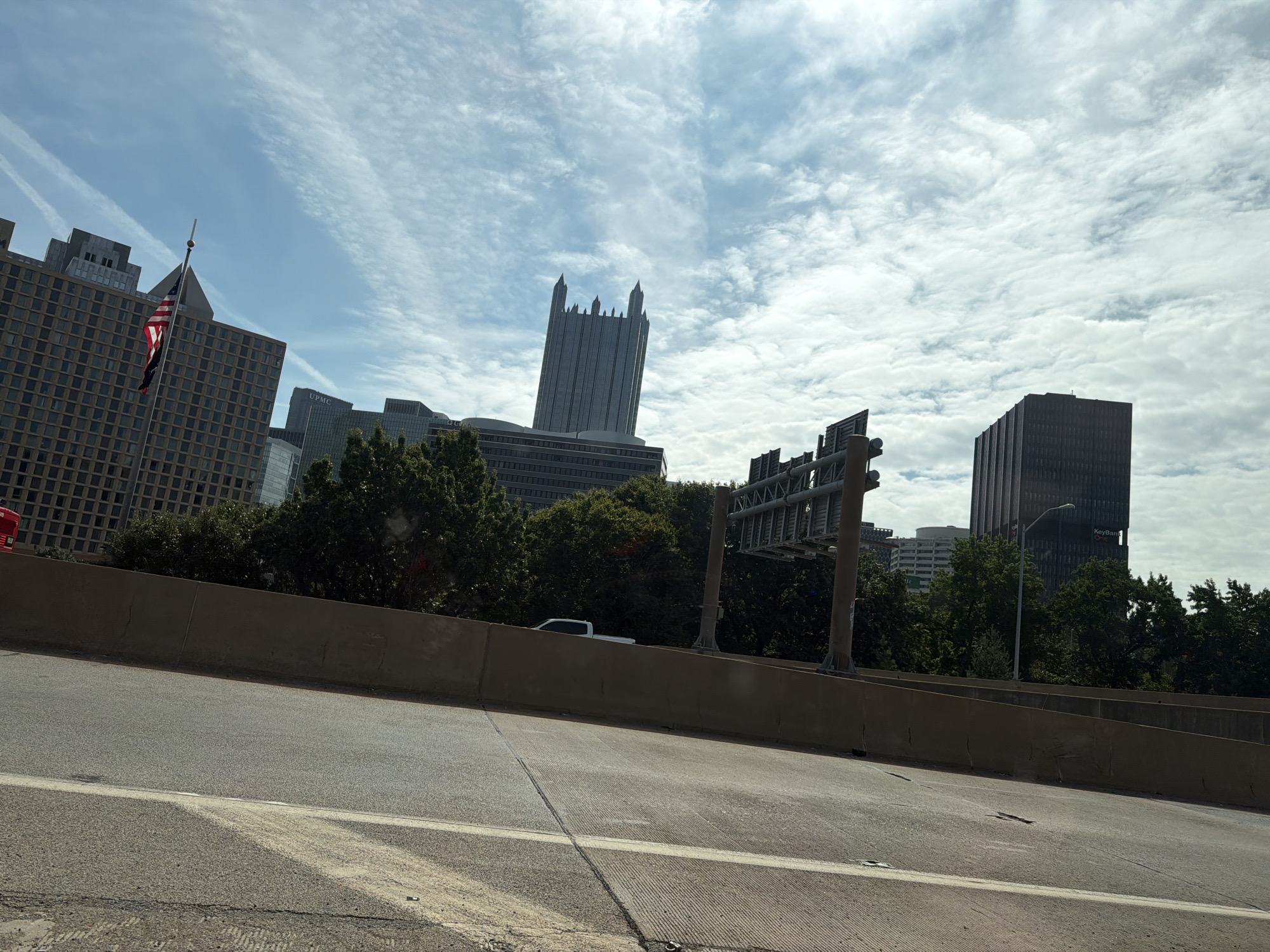 This is a medium-distance, eye-level, outdoor, daytime shot of the Pittsburgh skyline as seen from a highway overpass. The sky is partly cloudy with cirrus clouds streaking across the frame. In the foreground, a concrete barrier runs along the edge of the highway, with an asphalt road in front of it. In the midground, trees line the highway, partially obscuring several buildings. The most prominent buildings are the U.S. Steel Tower, a distinctive skyscraper with a crenellated top, and the Koppers Building, a dark, rectangular skyscraper. An American flag is visible flying from a flagpole in front of a brown, grid-patterned building. A white pickup truck is parked on the side of the road behind the concrete barrier. A highway sign is visible on a metal pole. The scene appears to be set in downtown Pittsburgh, Pennsylvania, United States.