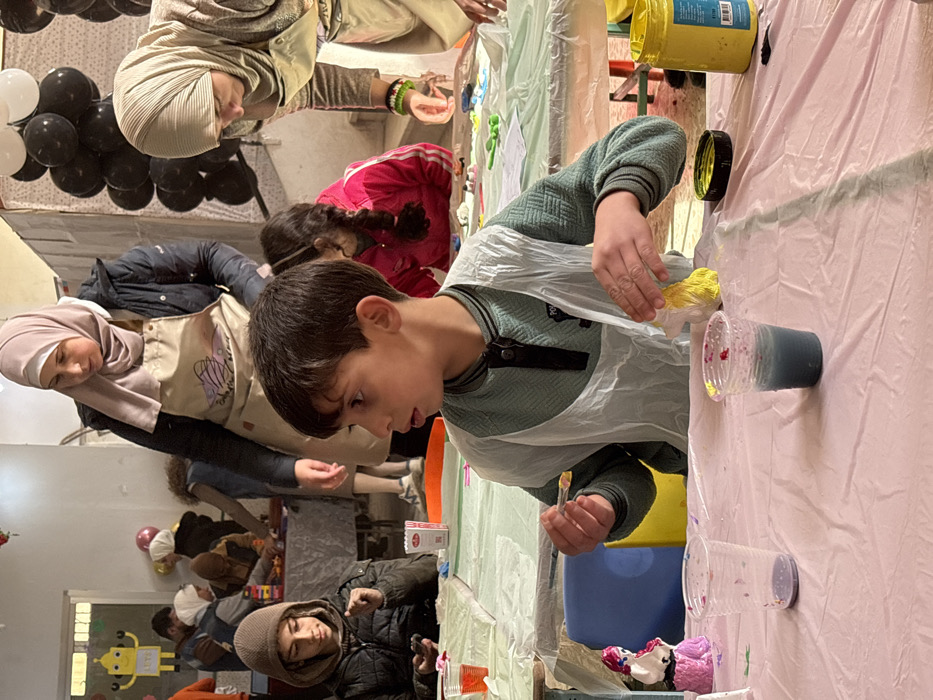 The image captures an indoor art and craft session occurring in Kafr Batna, Syria. In the foreground, a young boy with dark hair and fair skin is seated at a table covered with a pink plastic tablecloth. He wears a teal long-sleeved shirt and a translucent plastic apron. His head is turned to the left, and he holds a spoon containing a yellow substance in his right hand, while his left hand manipulates a yellow-colored, irregularly shaped object on the table. To his left on the table are two clear plastic cups; one is mostly empty with purple residue, and the other contains a blue liquid with red specks on its rim. A yellow paint jar with its black lid is also present on the table to his right. A white figurine with purple details stands on the table to his left.
Behind and to the left of the foreground boy, another boy wearing a brown hooded jacket is seated, looking down at a mobile phone in his hands. A clear plastic cup with red liquid and a small red and white box are on the table in front of him.
An adult woman, wearing a dark jacket, a light pink hijab, and a beige apron with a graphic design, stands to the left of the foreground boy, observing the activity. To the right, another adult woman, dressed in a light beige hijab and apron, leans forward over an adjacent table, engaged in a task; a bracelet displaying green, white, and red stripes is visible on her right wrist. A child in a pink jacket is partially visible behind the foreground boy, also participating in an activity.
The background reveals several other individuals, including adults and children, seated at tables covered with light-colored plastic sheets, all engaged in creative tasks. The room features light-colored walls. Decorations include a cluster of black and white balloons on the right and a yellow robot-shaped cutout with the letters "SET" displayed on the far-left wall.