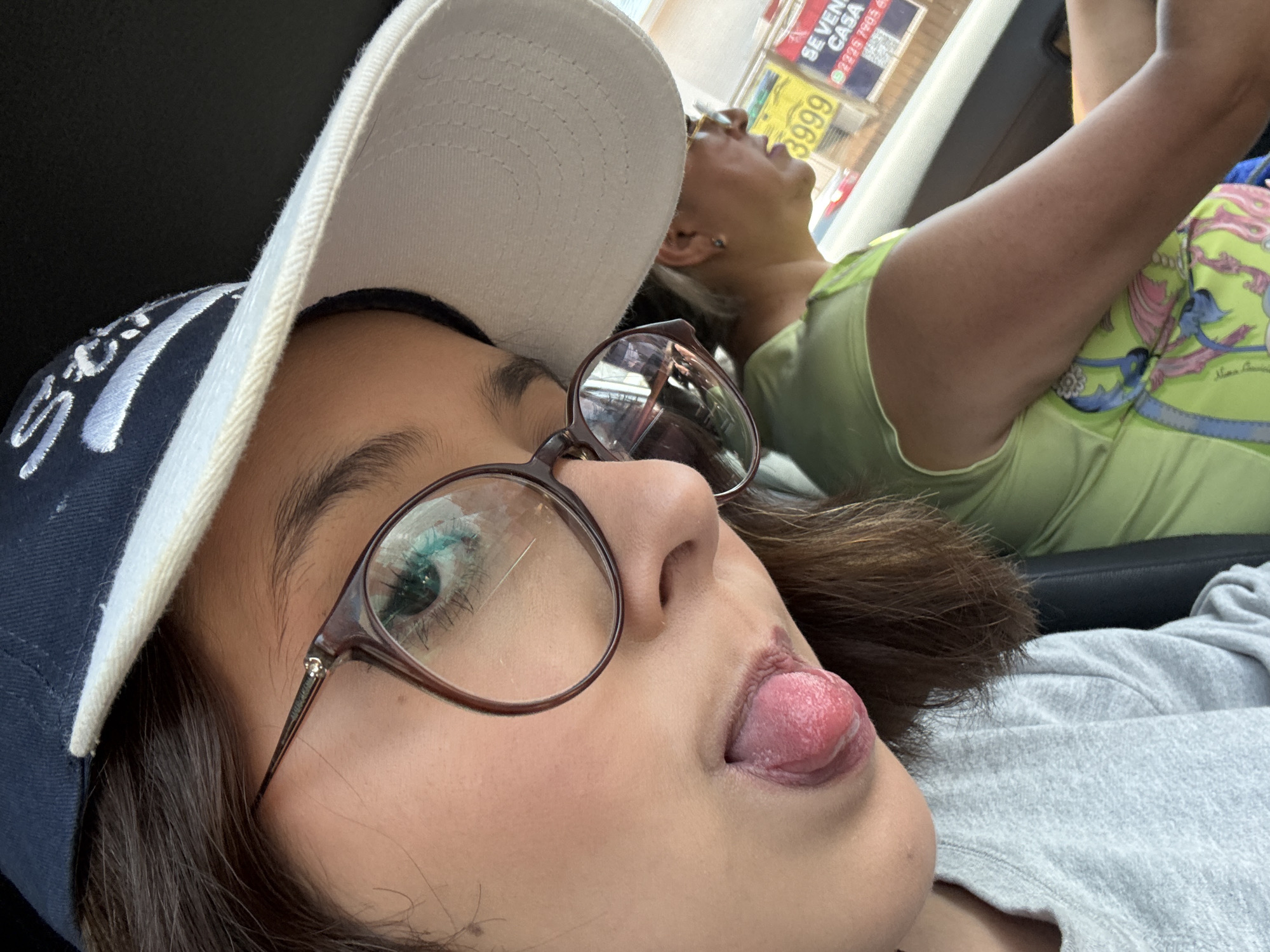 The image shows a close-up selfie of a young woman in the front passenger seat of a car. She is wearing a dark blue baseball cap with white embroidery, a gray long-sleeved shirt, and brown-rimmed glasses. Her tongue is sticking out. Another woman is visible in the background, leaning back in the driver's seat, wearing a green shirt. Outside the car window, there is a sign with the words "SE VENDE CASA" and a phone number. The sign indicates that they are in Puebla, Mexico.