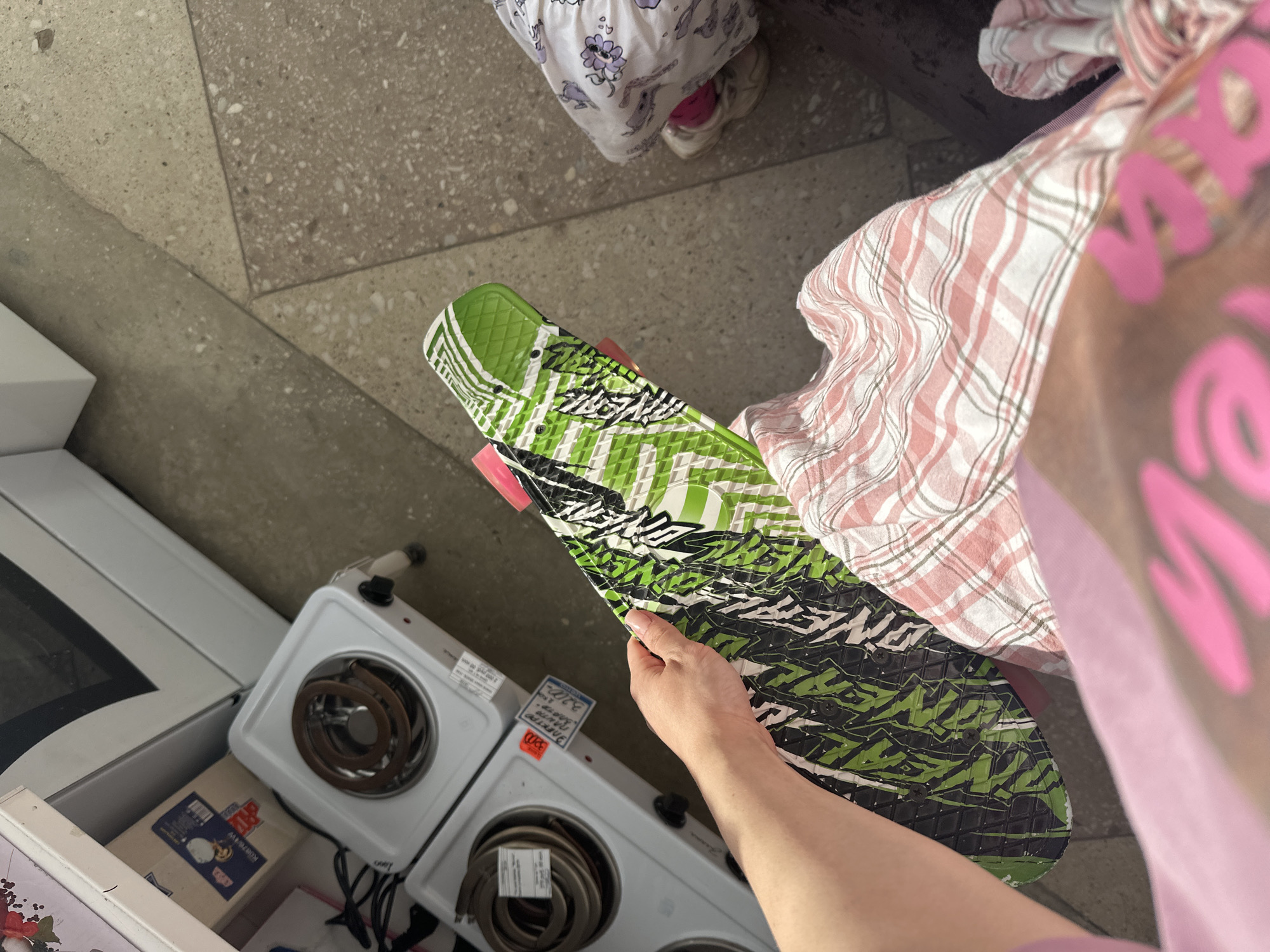 The image depicts a close-up, overhead view of a person holding a green and white patterned skateboard. The skateboard has a textured grip surface and pink accents on the wheels. The person's hand, with manicured nails, is visible grasping the board. In the background, two portable electric stovetops are present, one on top of the other, suggesting a retail or storage environment. A price tag with Russian text is affixed to the lower stovetop. The floor appears to be a mix of concrete and tile. To the right of the skateboard, a person wearing pink socks and white sneakers is partially visible, along with patterned fabric, possibly clothing or a blanket. The overall scene suggests an indoor location, possibly a store or apartment, with domestic appliances and a recreational item. The specific location of Yaroslavka, Russia is not directly evident from the visual information in the image itself.