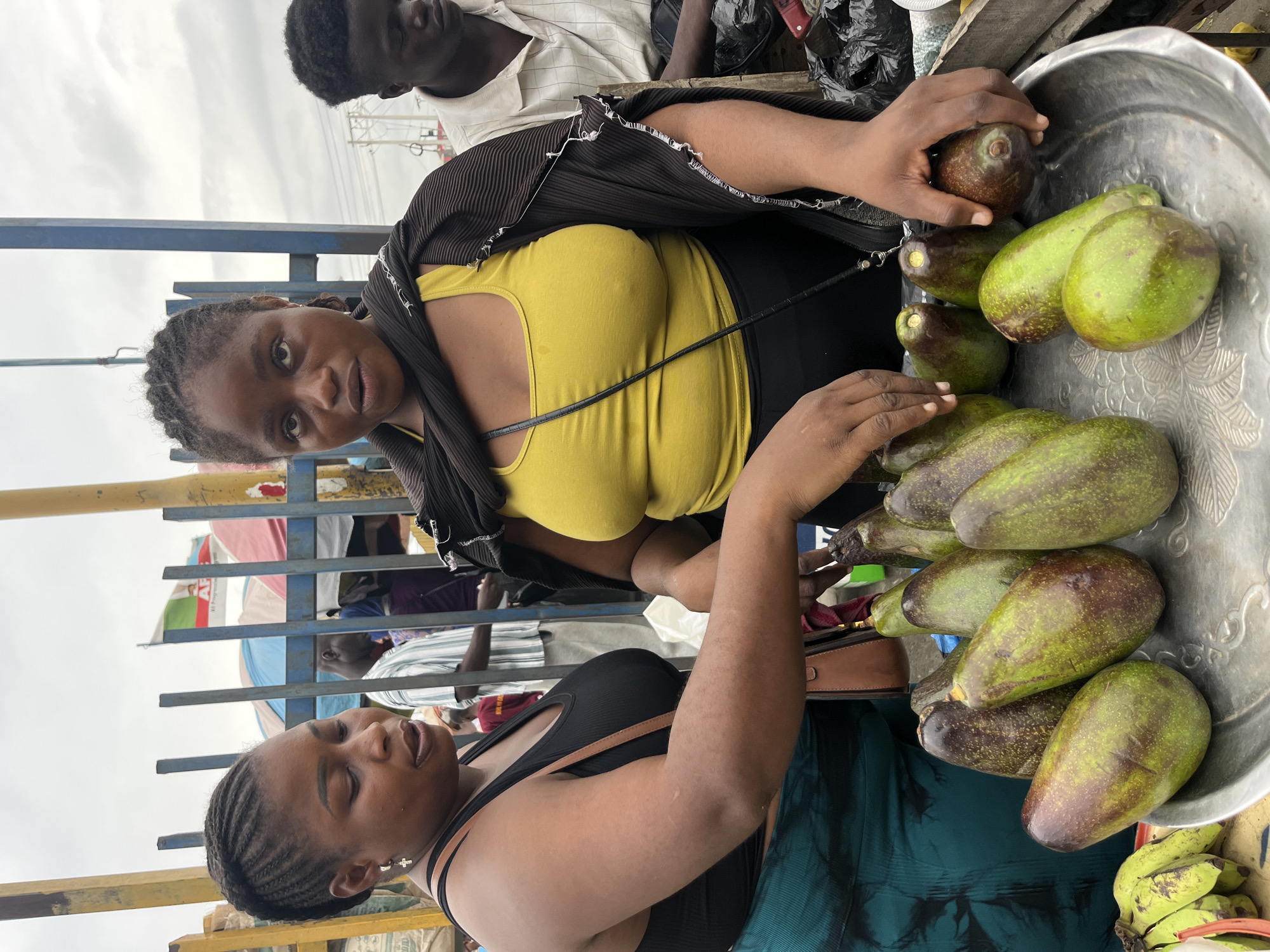 Two women are examining avocados in Lekki, Nigeria. One woman, wearing a yellow top and a striped shawl, holds an avocado in her hand, looking directly at the camera. The second woman, in a black tank top, is also inspecting avocados from a large silver bowl. The avocados vary in color from green to brownish-purple. Additional avocados and some bananas can be seen next to the bowl. Other individuals are visible in the background near a fenced area.