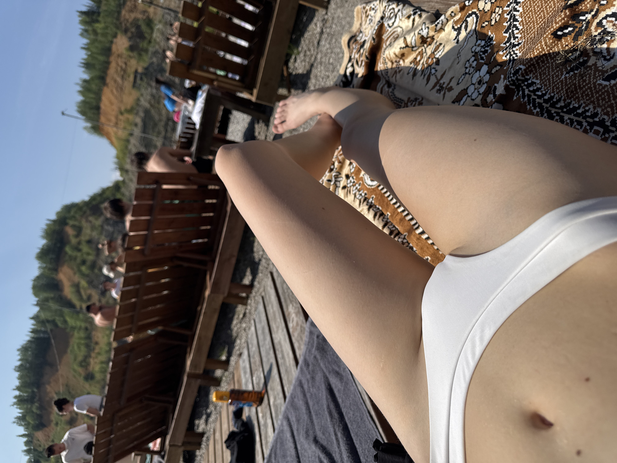 The image shows a person lying on a lounge chair with a patterned blanket under them. The person is wearing white bikini bottoms. Their legs are visible, and one foot is resting on the blanket. In the background, there are more lounge chairs, a table, and other people sunbathing and relaxing. There are also trees and a hillside in the background. The location is described as Karyer Izvestnyak, Russia.