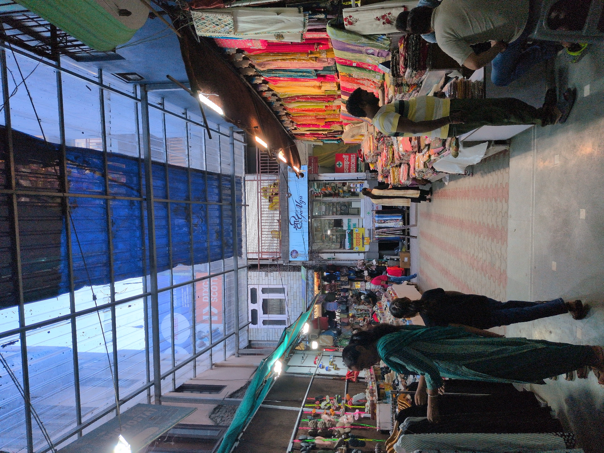 The image depicts a busy market street in Kullu, India, covered by a transparent and blue corrugated roof, supported by metal beams. The market stalls display a variety of colorful clothing and accessories. On the left side, a woman in a teal kurta is examining garments on a rack, accompanied by a young girl in a black shirt and blue pants. Further down the street, various shops are visible, including one with mannequins adorned in traditional Indian attire. On the right side, a man in a striped yellow and white shirt is standing next to a stall piled with folded fabrics. Another man in a grey shirt is kneeling in the foreground, facing away from the camera. The street is paved with a patterned tile walkway. The atmosphere suggests a lively commercial environment.