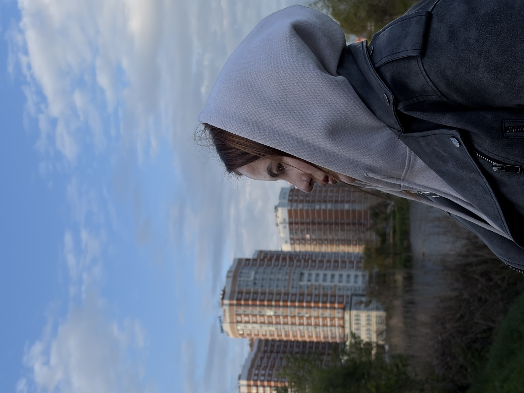 The image captures a young person in profile, standing outdoors in an urban setting under a partly cloudy sky. The individual, likely female, is positioned on the right side of the frame, wearing a light grey hooded sweatshirt with the hood pulled up, and a dark grey or black jacket over it. Her brown hair is visible, and her gaze is directed downwards, conveying a contemplative or pensive mood.
In the mid-ground, several multi-story residential buildings with red-brown and white facades are visible, characteristic of modern apartment blocks. These structures are somewhat blurred, suggesting a focus on the foreground subject. The lower left portion of the image shows blurred greenery, possibly trees or bushes, indicating a park-like area within the city.
The sky, occupying a significant portion of the image, is a clear blue with scattered white and light grey clouds, suggesting fair weather. The soft, natural lighting points to daytime, possibly late afternoon. There is no specific activity depicted beyond the person's presence in the scene. No text is visible. The scene, with its blend of modern urban architecture and green spaces, aligns with the landscape found in cities such as Krasnodar, Russia.