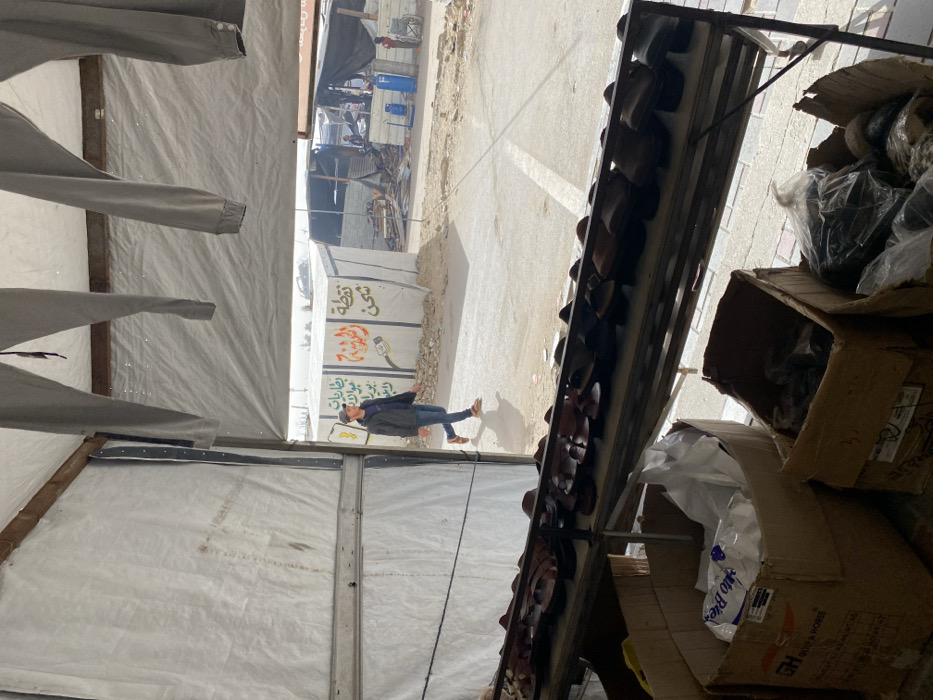 A street scene viewed through a tent-like structure. Inside, clothes hang on a makeshift rack, while outside, a man in jeans and a jacket walks on the side of the road. A cart displays items, and boxes sit nearby. A building with graffiti and advertisements is visible, with people in the distance.