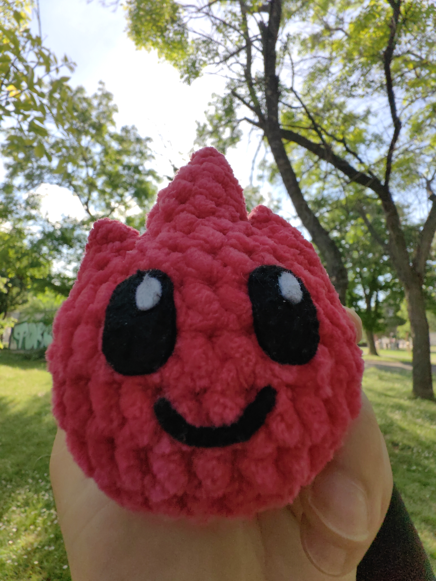A red crocheted plush toy with black eyes and a curved black mouth is being held in a person's hand. The background is a public park with green grass and trees with green leaves. Graffiti is visible on a wall in the background. The location is likely Shumen, Bulgaria.