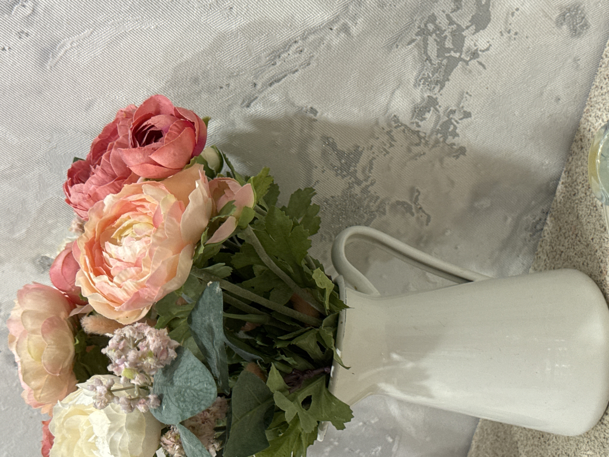A still-life photograph features a bouquet of artificial flowers in a white pitcher. The flowers are predominantly pink and peach, with some white and green foliage. The pitcher is tilted to the side. The background is a textured, greyish-white surface, possibly a wall. A portion of a light-colored fabric is visible in the lower right corner of the frame. No people are present. No geolocation information can be inferred from the visual data.