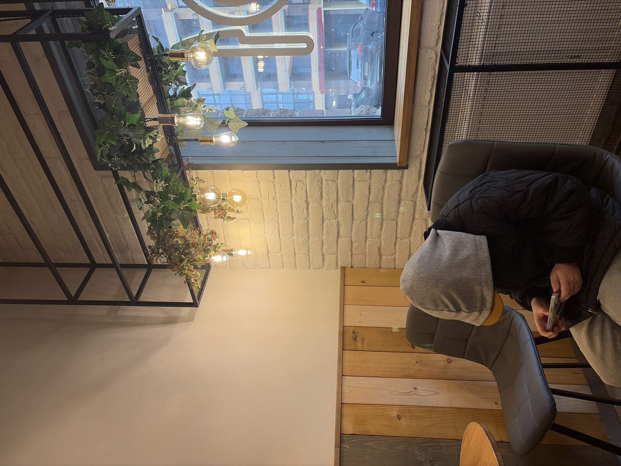 A person in a grey hooded sweatshirt and black jacket sits in a grey cushioned chair, holding a phone. The scene is inside a building with a window showing a street view, including cars and buildings, suggesting a city environment. The interior features a white brick wall, a light-colored wall, and a wooden wall paneling. Above, a black metal structure holds plants and light bulbs. Based on available visual indicators such as architectural style and car models, the location is likely Saint Petersburg, Russia.
