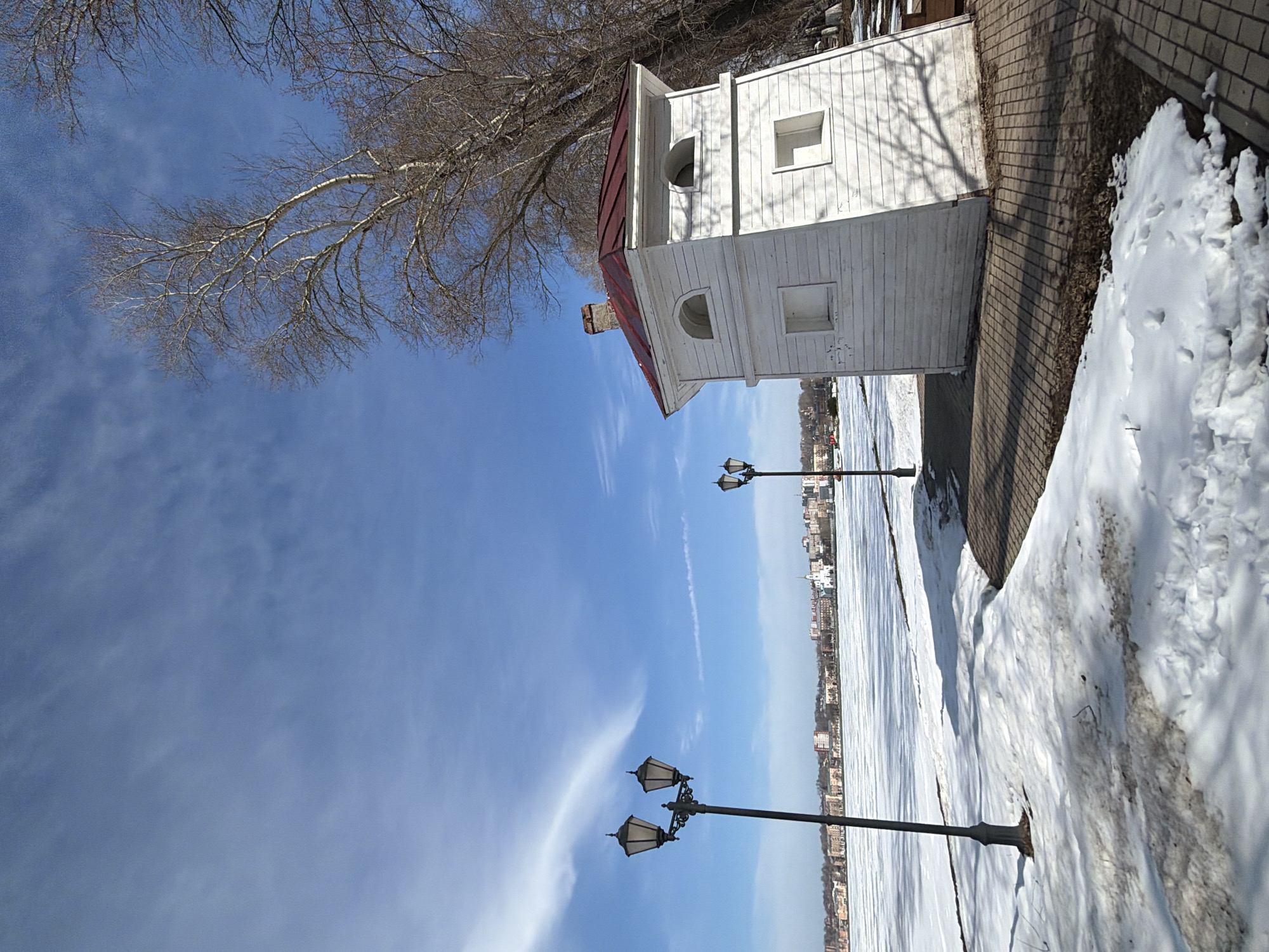 The image depicts a winter scene in Votkinsk, Russia, featuring a small, white lighthouse-style structure with a red roof. It is positioned on a paved embankment, alongside a snow-covered ground area. Two ornate streetlights stand along the embankment, against the backdrop of a frozen body of water (likely a river or lake) and the city skyline in the distance. The sky is mostly blue with some scattered clouds, and bare tree branches are visible in the upper part of the image.