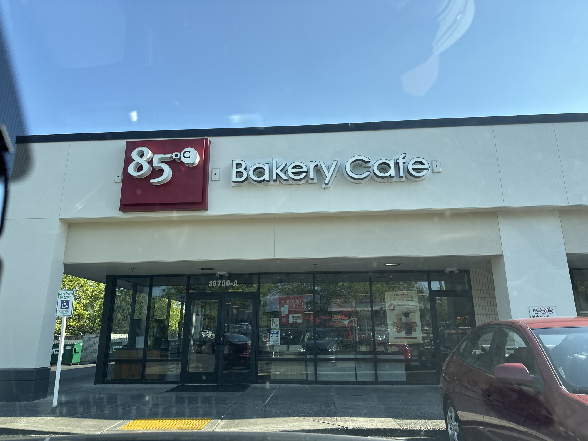 The image shows the exterior of a "85°C Bakery Cafe" located at 18700-A in Lynnwood, United States of America. The building has a large glass storefront with the company's logo and name prominently displayed above the entrance. The signage features "85°C" in a red square with white lettering and "Bakery Cafe" in white, illuminated letters. To the left of the entrance, there is a reserved parking sign with a wheelchair symbol. Inside the cafe, through the glass, shelves stocked with baked goods and a counter area are visible. Several cars are parked in front of the building and in the background. The sky above is clear blue with some sunlight reflecting on the windows. The overall scene depicts a commercial establishment on a sunny day.