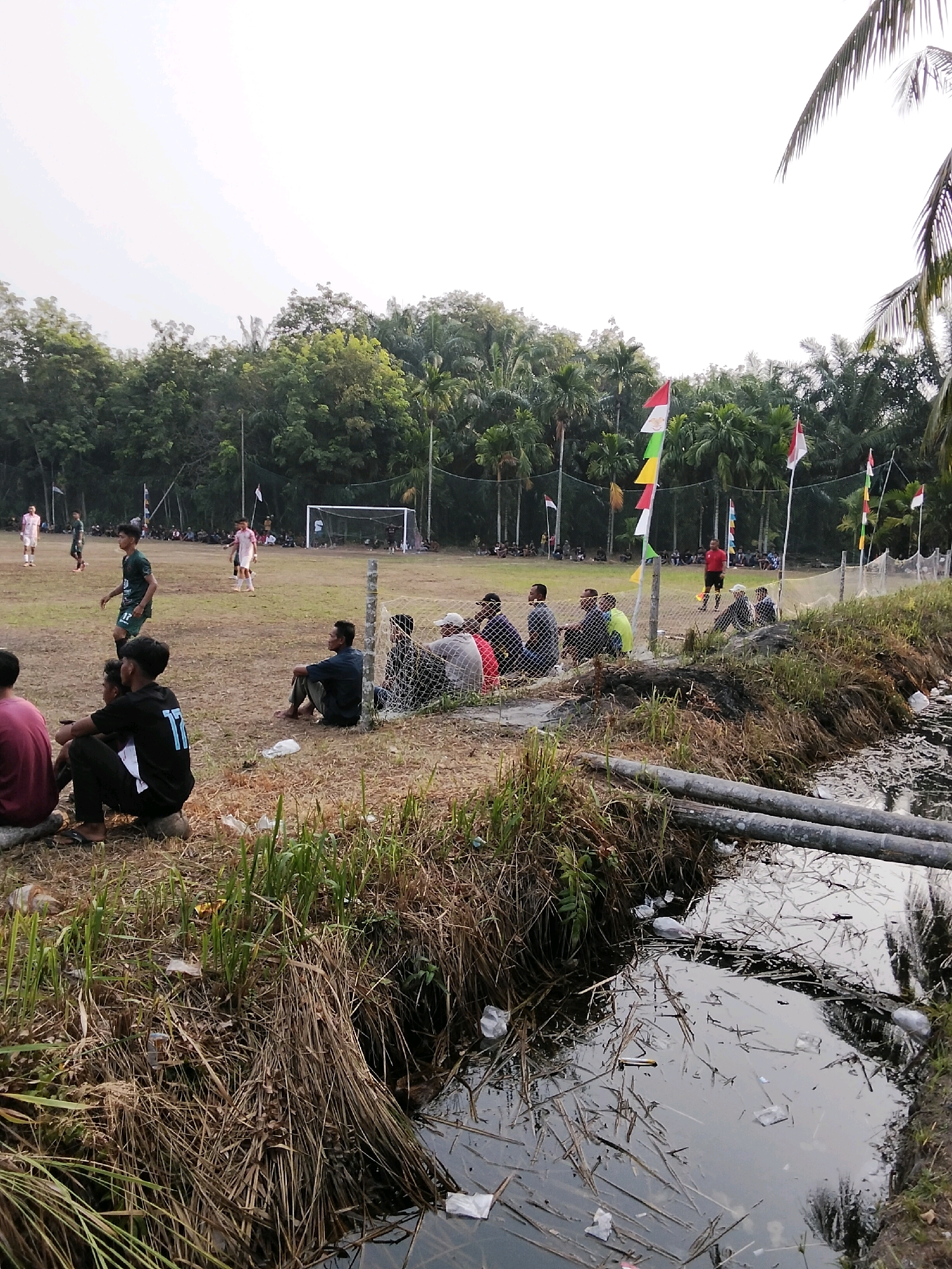 In Pambang Pesisir, Indonesia, a rural football match is underway on a dirt field lined by a fence. Spectators, some seated on the ground, others on rocks, watch the game from behind the fence. In the foreground, a small stream flows with debris, including plastic bags and dried vegetation. Beyond the field, a line of trees forms the backdrop, with palm trees interspersed among them. Indonesian flags and colorful triangular pennants are visible, adding a festive touch to the scene. The sky is overcast, suggesting a cloudy day.