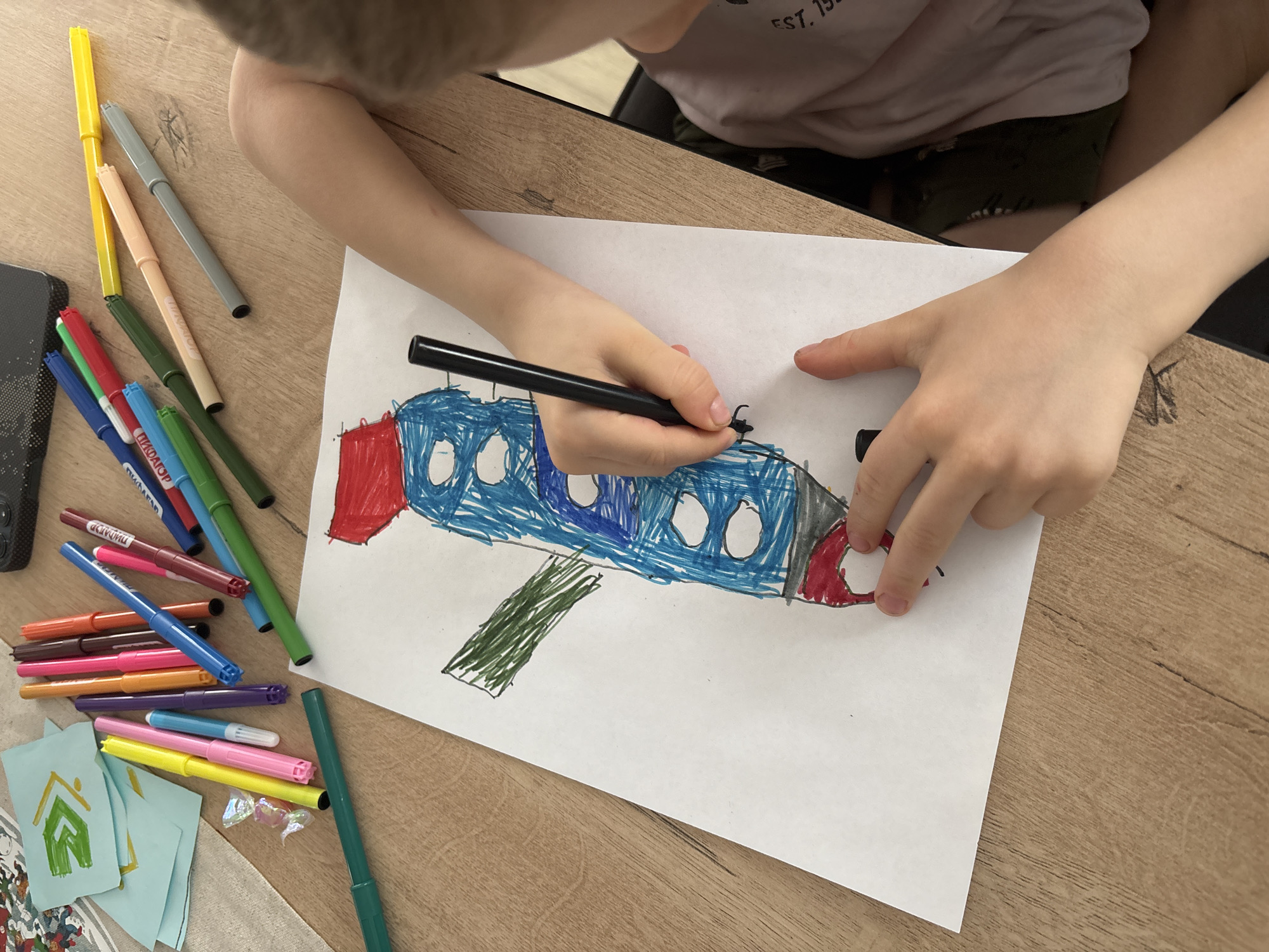 A child is drawing an airplane on a sheet of white paper with a black marker. The airplane is colored with blue, red, and green markers. Various colored markers are scattered around the drawing on a wooden table. A few small, light blue cards with a green house-like drawing are also present. The scene appears to be indoors. The location of Chaikovsky, Russia cannot be determined from the image.