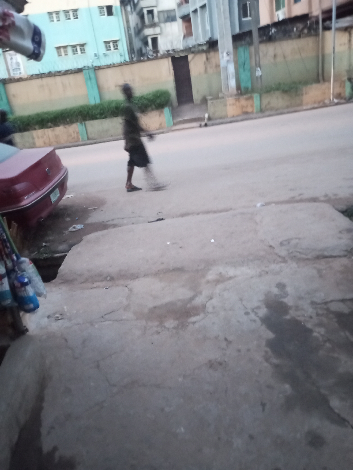 This image depicts an outdoor street scene in Onitsha, Nigeria, captured during either early morning or late afternoon, as suggested by the soft, diffused light.
In the middle ground, a blurred figure of a person, dressed in dark clothing, is visible moving across the asphalt road from right to left. They appear to be performing an activity like sweeping, with their right arm extended forward.
To the left of the frame, the rear end of a red car is partially visible, parked or pulled over beside the road. The car has a green license plate, and faint lettering on its trunk identifies it as a "PEUGEOT." Between the car and the concrete sidewalk, there's a visible ditch or drainage channel, containing some litter. In the far left foreground, several items, possibly cleaning implements like brooms or mops, are hanging, suggesting a nearby stall or shop.
The background features several multi-story buildings. A prominent light blue building with numerous windows stands behind a long wall, which is painted in alternating sections of light green and beige and topped with a fence. Other buildings, varying in shades of grey and beige, are also visible. The street and sidewalk surfaces are made of concrete and asphalt, showing some wear and tear, and there is scattered debris on the ground. The overall scene portrays a bustling urban environment.