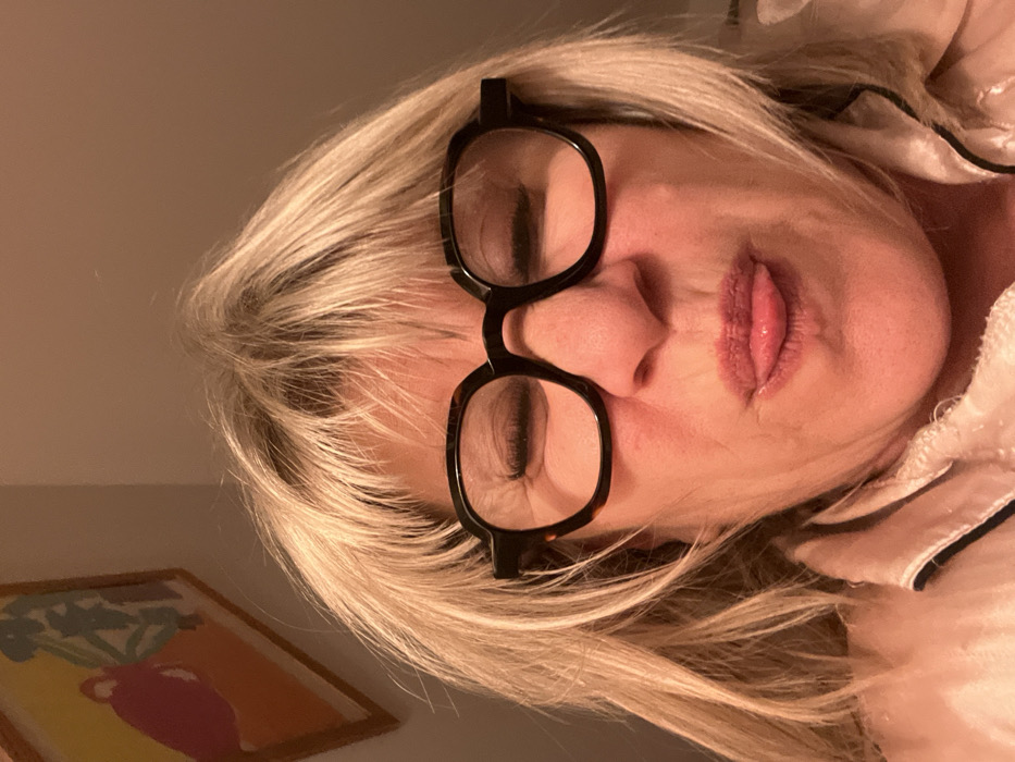 A woman with blonde hair and black-rimmed glasses is seen with her eyes closed and tongue slightly protruding. Her skin tone appears fair, and she wears a satin-like top. A framed picture is faintly visible in the lower left corner. The image is slightly tilted.