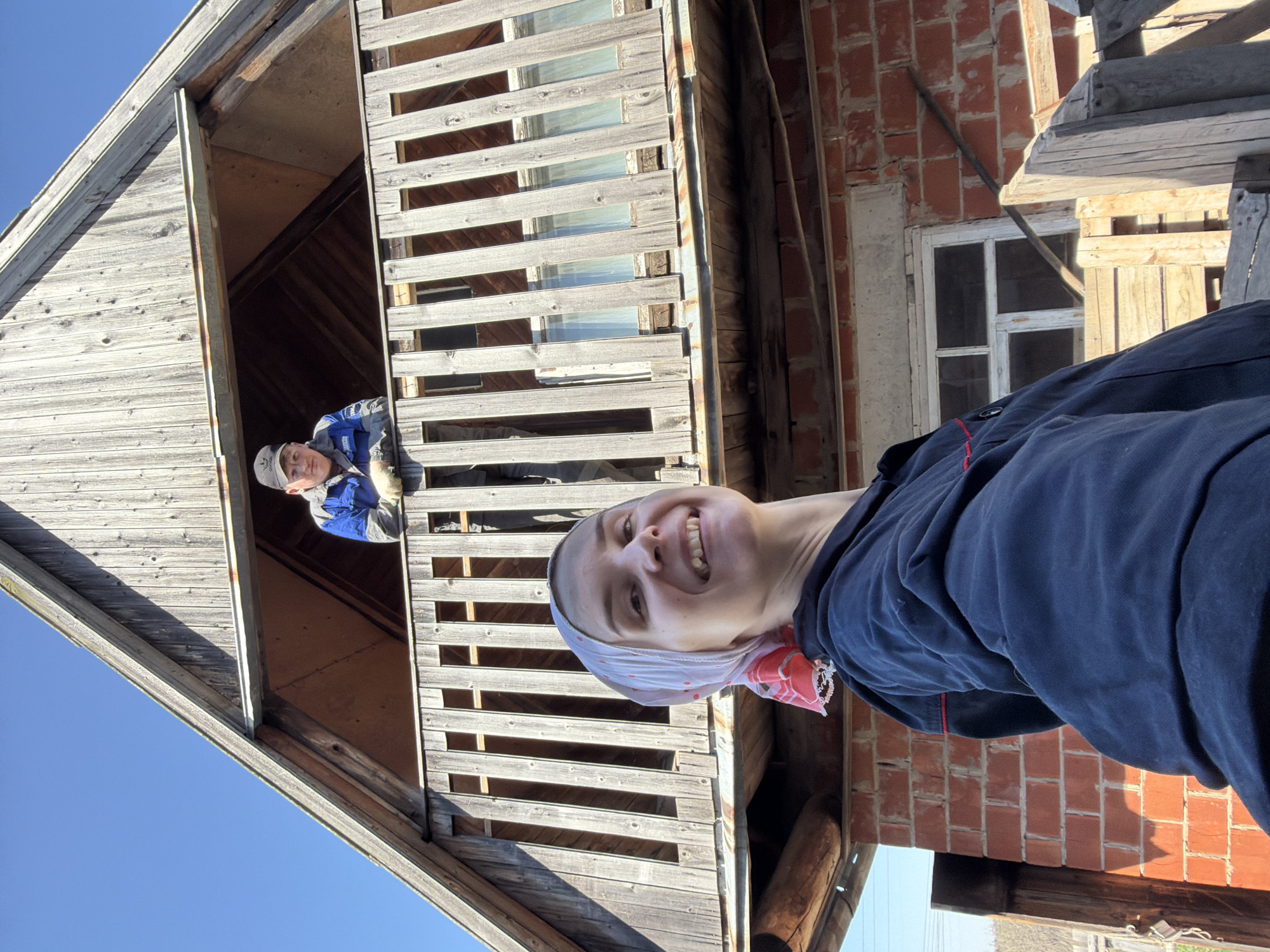 A low-angle photograph shows two individuals in front of a wooden and brick structure under a clear blue sky. The location is Chaikovsky, Russia.
A person with a headscarf and a dark blue jacket is positioned in the foreground, facing the camera with a smile. Behind this person is a red brick wall with a white-framed window. To the left, a weathered wooden structure stands with a peaked roof.
On the upper level of the wooden structure, visible through an opening, stands another person wearing a hat and a blue jacket. The wooden structure has open-slatted windows and a balcony-like area. The lighting indicates it is daytime.
