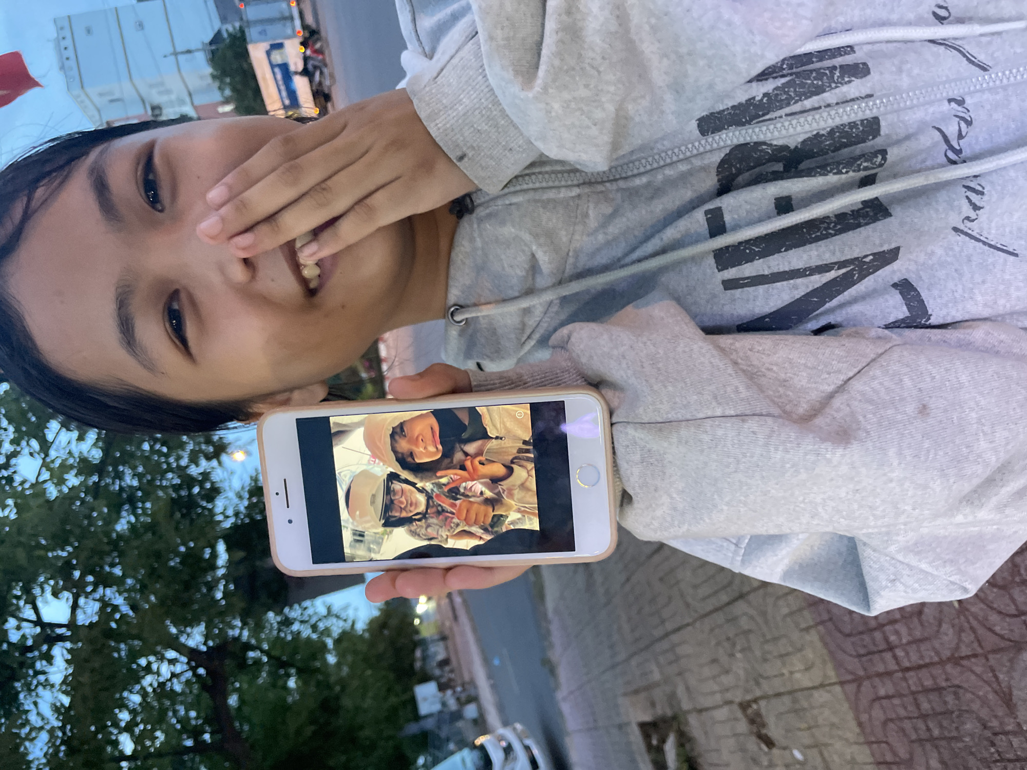 A young girl is holding up a smartphone displaying a selfie of herself and another person. Both individuals in the selfie are wearing helmets and appear to be on a motorcycle, with one giving a thumbs-up and the other making a peace sign. The girl in the foreground is smiling and covering her mouth with her hand. She is wearing a grey hoodie with a graphic print on the front. The background suggests an outdoor urban environment in Mũi Né, Vietnam, with trees, a street, buildings, and vehicles visible. The lighting indicates it is either early morning or late evening.