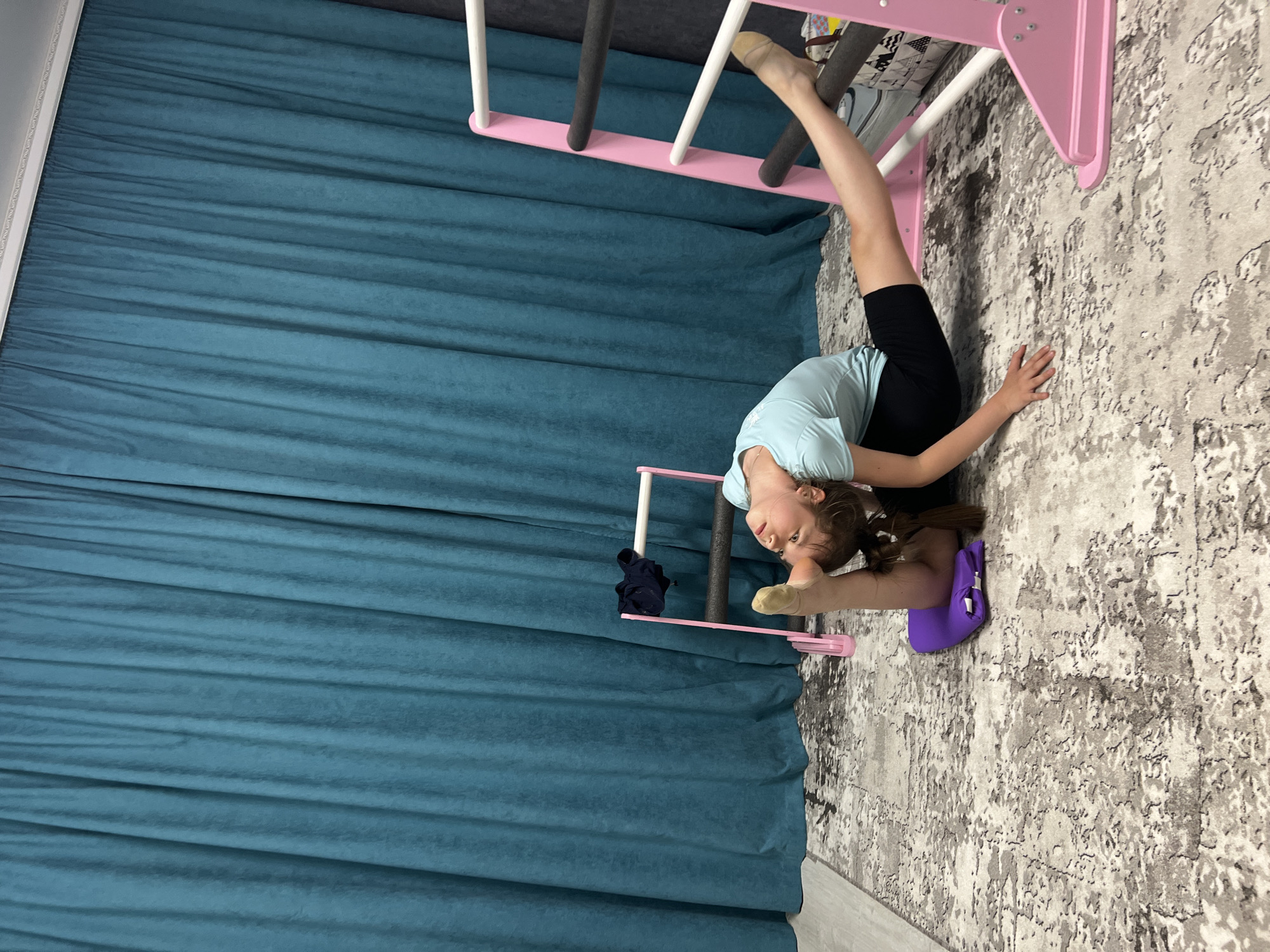 A young girl is performing a gymnastic pose indoors. She is positioned on a patterned carpet with a purple cushion beneath her lower back for support. Her left leg is elevated, bent at the knee, and resting on a horizontal bar of a pink and white climbing frame. Her right leg is extended forward, almost perpendicular to her torso. She wears a light blue short-sleeved shirt and black shorts. Her hair is pulled back in a ponytail. A blue curtain is visible in the background, covering a significant portion of the image. A pink gymnastic wall ladder with black pads is also visible. The image's location is in a room in the city of Chaikovsky, Russia.
