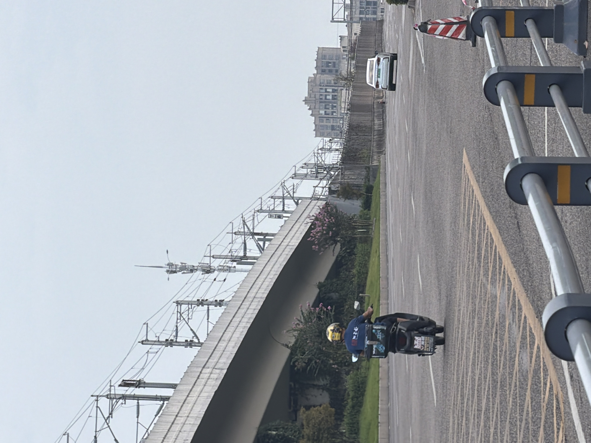 In Jiangjin, China, an overcast day unfolds on a multi-lane road. A person, wearing a dark blue shirt and a yellow helmet, rides a black electric scooter from left to right. The scooter is equipped with a large, black delivery box on the back, adorned with a blue and white logo. The rider appears to be on the left side of the road, moving away from the viewer.
To the left of the road, an elevated concrete structure, likely a bridge or overpass, dominates the landscape. Overhead, a complex network of electrical wires and support poles, characteristic of a railway or power line system, crisscrosses the sky. Lush green vegetation and some flowering bushes with pink blossoms grow beneath the elevated structure.
Further down the road, a white car is traveling in the same direction as the scooter. In the background, a multi-story building with a distinct architectural style stands against the hazy sky. Traffic cones, one with red and white stripes, are placed along the side of the road, suggesting ongoing construction or lane closures. A metal guardrail with yellow markings is visible in the foreground, separating the roadway from the viewer. The road surface is asphalt, with visible lane markings.