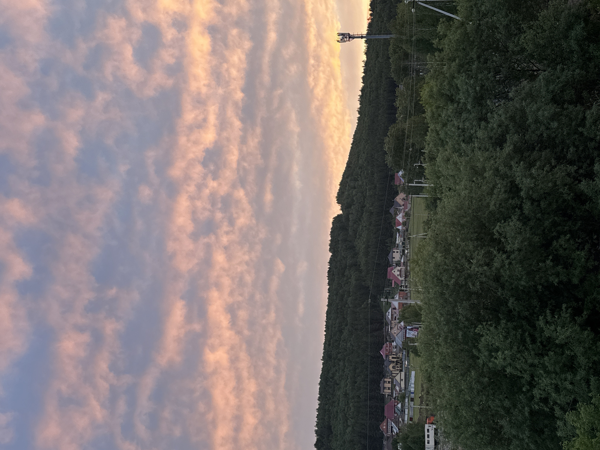 The image depicts a landscape with a sky filled with long, streaky clouds that are tinged with pink and gold, suggesting either sunrise or sunset. To the right of the sky is a dense green forest that appears to grow along a hillside or mountain. Along the base of the forest, there is a small town or village with houses that have distinct rooftops, some colored red. There are also power lines and a communication tower visible in the upper right of the image. The image is taken from a slightly elevated perspective. The location is in the city of Unknown, Unknown.