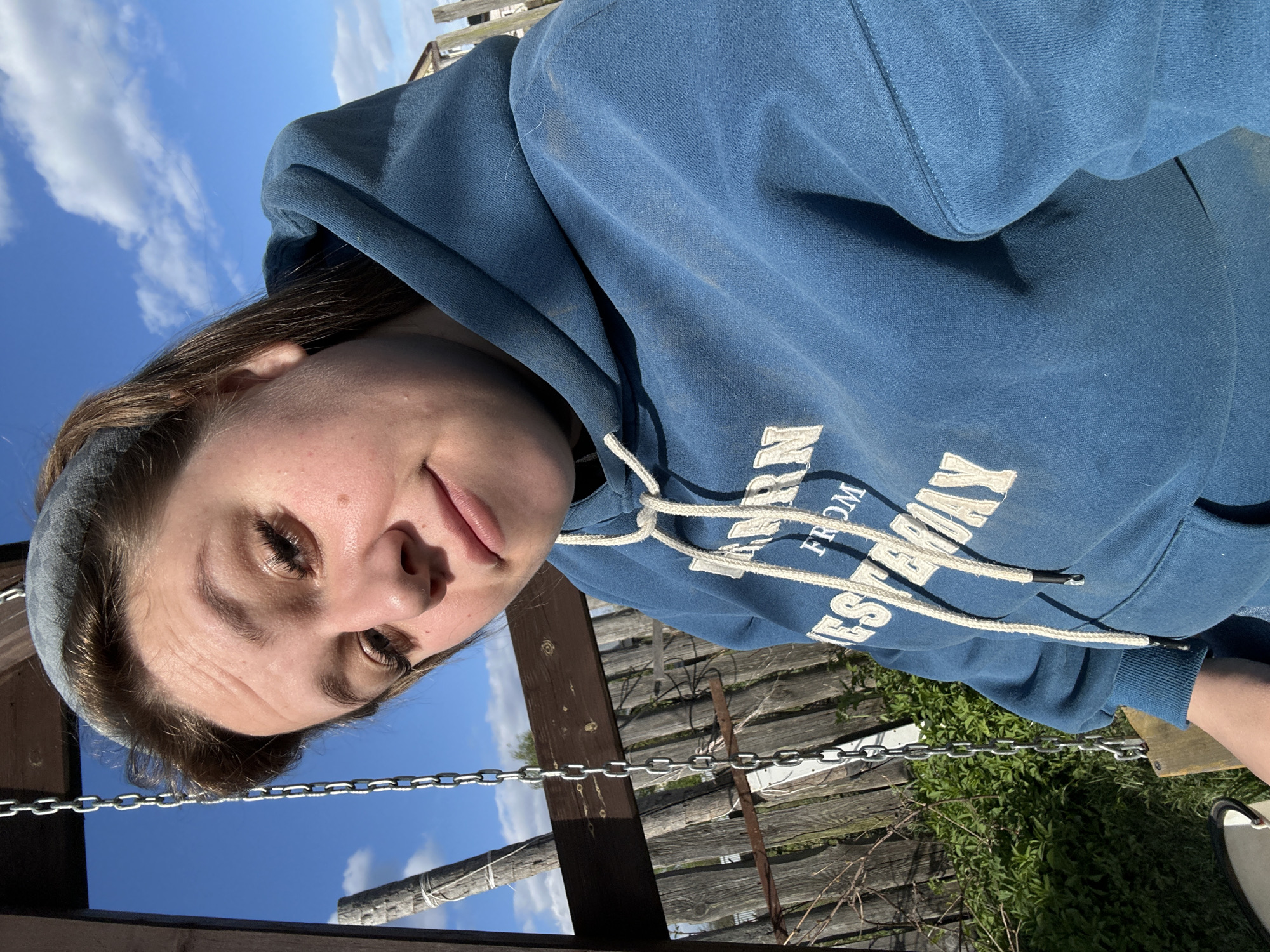 A woman in a blue hooded sweatshirt is the primary subject of this photograph. She is positioned in the foreground, and her face is in focus. Behind her, there's a wooden fence and some greenery, with a bright blue sky and white clouds visible above. A metal chain and part of what appears to be a swing set are also visible in the middle ground. The location is identified as Ольховка, Russia.