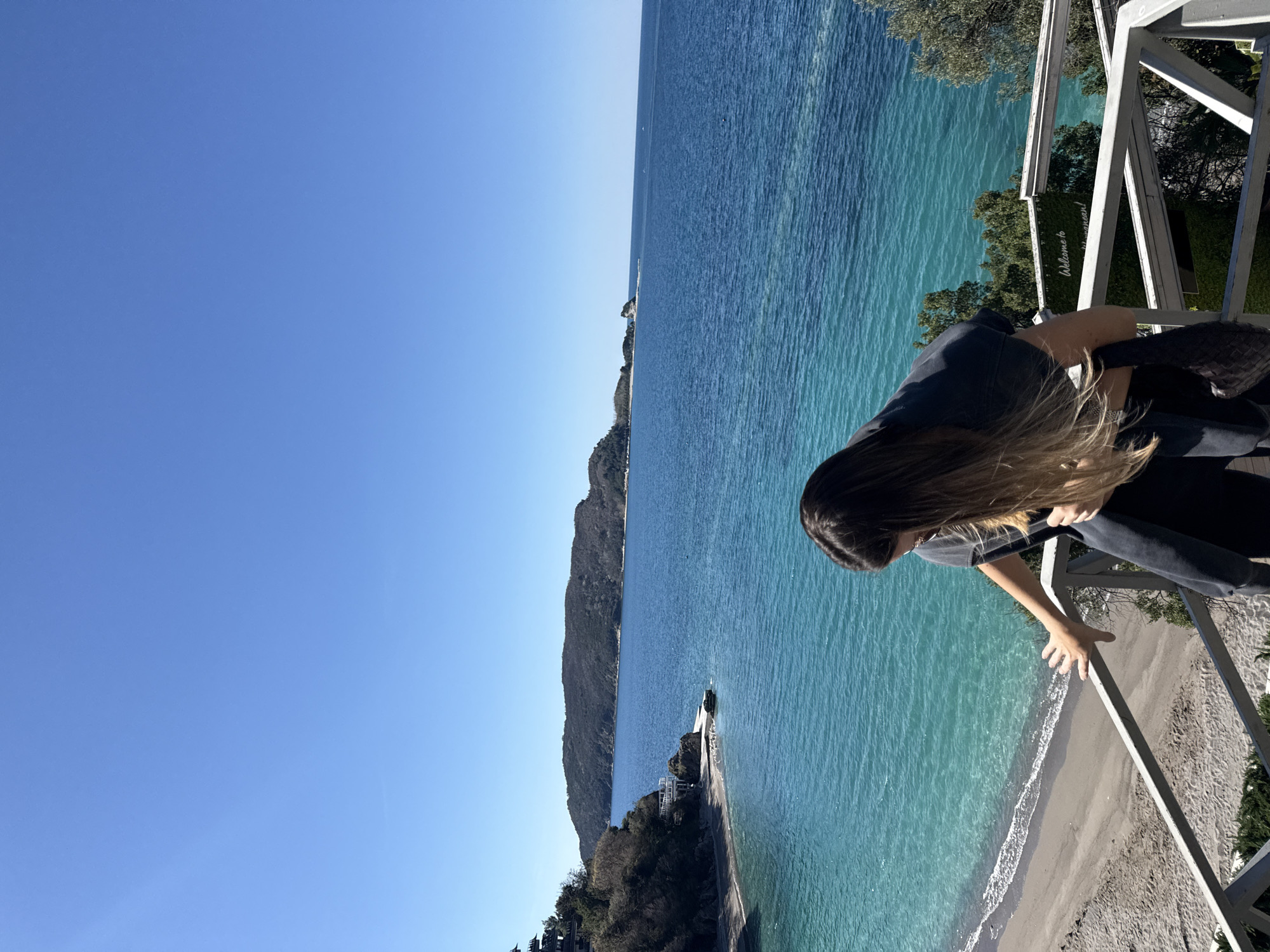 The image depicts a woman standing on a wooden balcony overlooking a coastline. The woman has long brown hair and is wearing a dark shirt and jeans. She is leaning over the railing, with one hand resting on it. The coastline features a beach with light sand and clear turquoise water. A small peninsula extends into the sea, partially covered with trees. In the background, a mountain or large hill is visible. The sky is clear blue. The location appears to be coastal, possibly a resort or scenic viewpoint. The city is unknown.