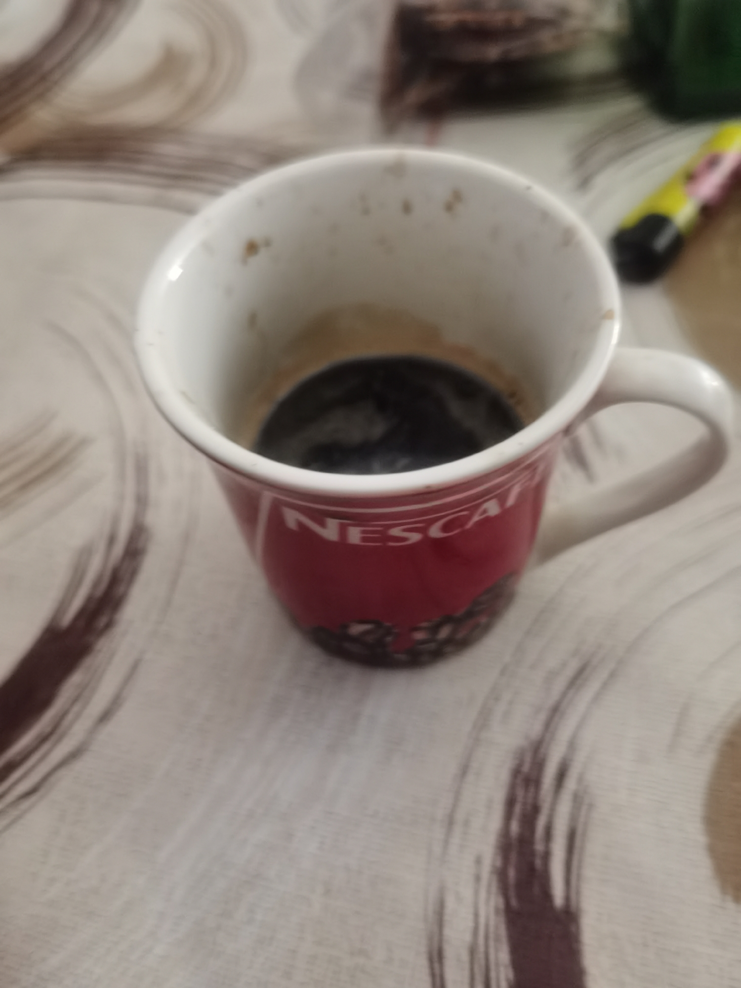 A close-up shot captures a red Nescafe mug, partially filled with dark coffee, resting on a patterned tablecloth. The mug has a white rim and handle. To the right of the mug, a yellow and pink object, possibly a marker or pen, is partially visible. The background is blurred, showing more of the tablecloth and some indistinct items. The scene appears to be indoors, likely on a table. There are no people present. The image was taken in Lomets, Bulgaria.