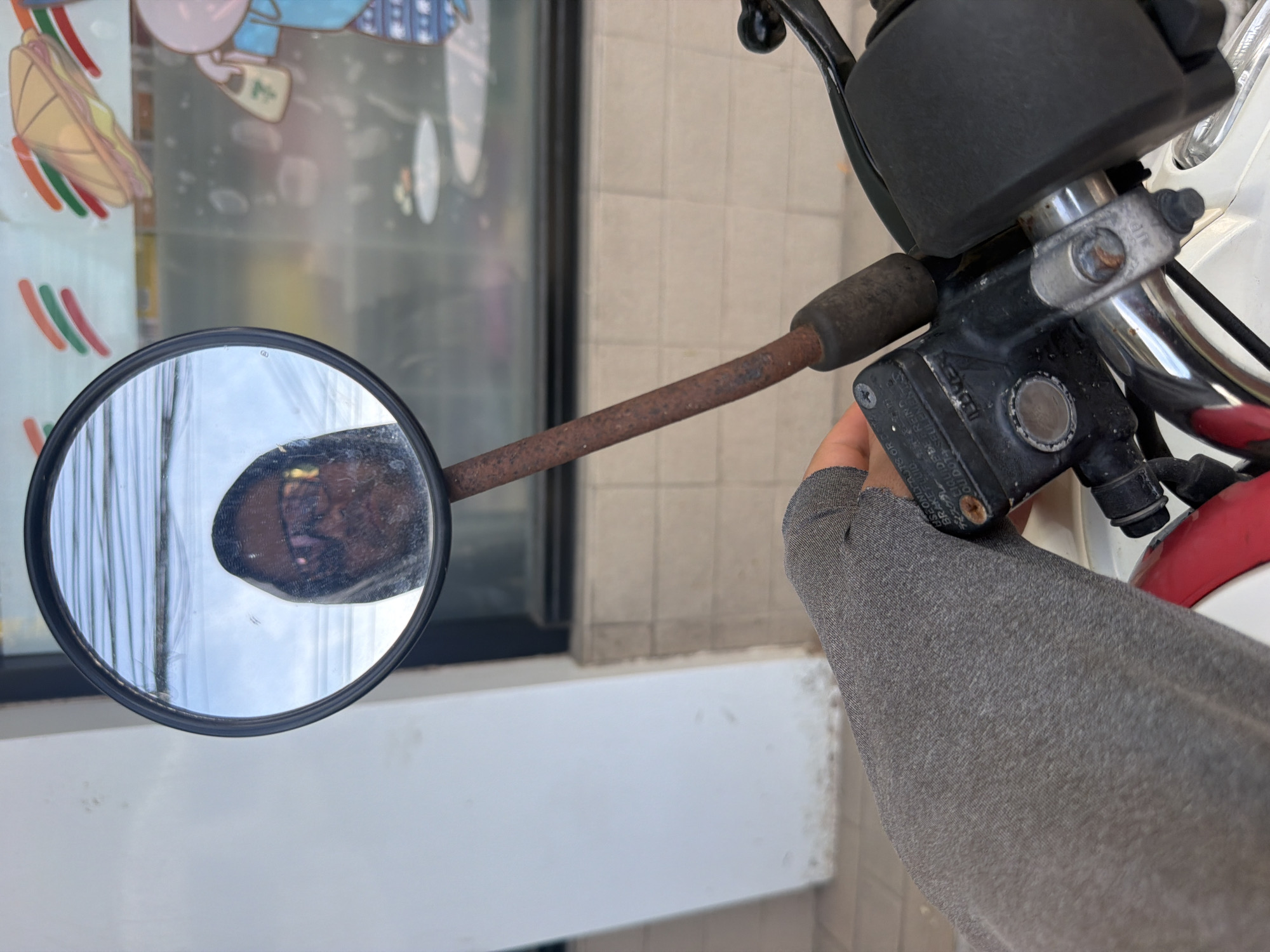 In Baan Mae Nam, Thailand, the image captures a close-up perspective from the viewpoint of a motorcycle rider. A gray sleeve covers the rider's left arm and hand, which is positioned on the motorcycle's handlebars, near the brake lever. A black plastic brake fluid reservoir is visible, with some text on it, including the word "LIQUID." The rusty stem of a circular rearview mirror extends upwards, reflecting a person wearing sunglasses and a hooded garment. The background shows a storefront with a white tiled facade and a glass window displaying signage with red, green, and orange stripes, and an illustration of a sandwich. The scene appears to be an outdoor urban environment.