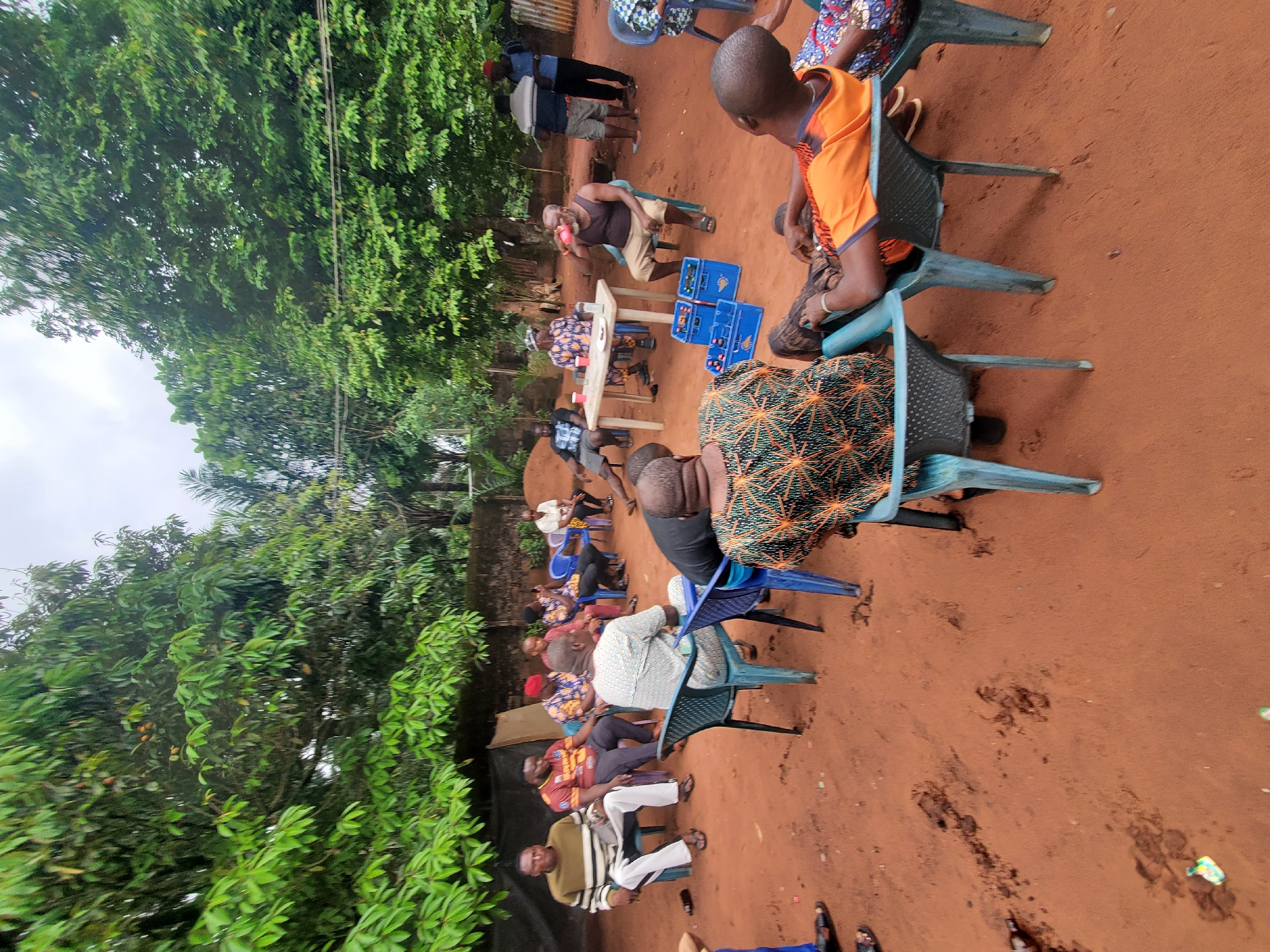 A gathering of approximately 15 people is taking place in Nnobi, Nigeria. The setting appears to be an outdoor space with red soil ground, with a lush green tree line framing the scene on the left. Individuals are seated on plastic chairs arranged in a somewhat informal manner. A small table holds what appear to be beverages and other objects. Several blue crates are located near the table. Participants are of varying ages and dressed in casual clothing. The weather seems overcast.