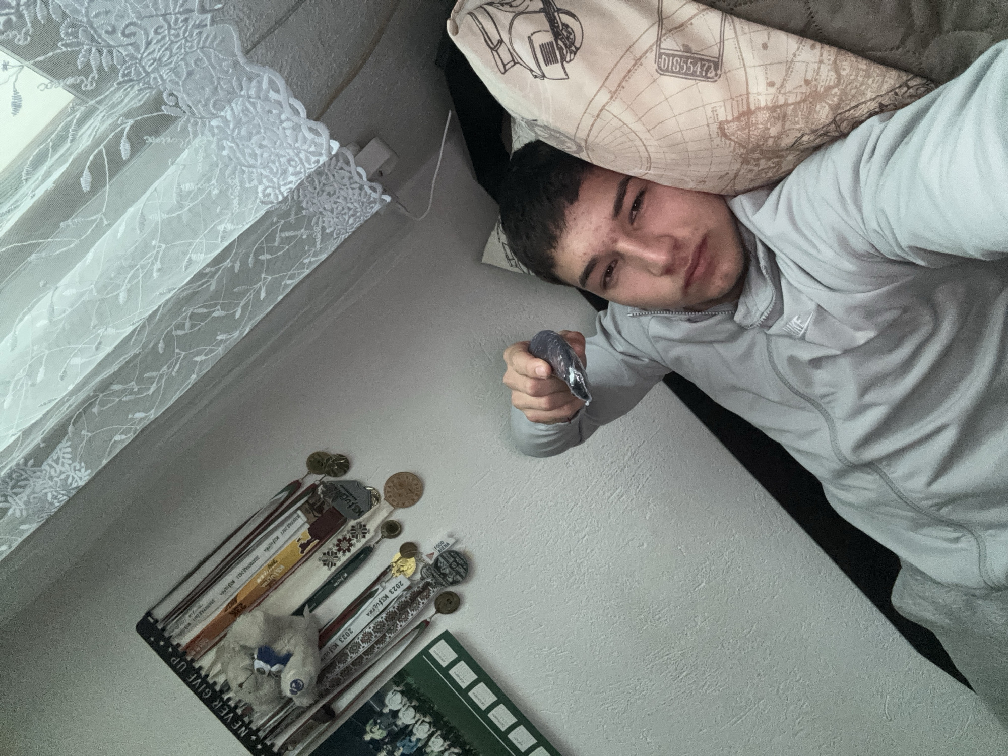 A young man is lying on a bed with a pillow under his head, holding a small, metallic object in his outstretched hand. He is wearing a grey athletic jacket with a visible Nike logo. To the left of the man, a window is covered with a white, lace-patterned curtain. Below the window, on a shelf mounted on the wall, there are several books, a small stuffed animal with a blue bow, coins, and medals. Inscriptions in Cyrillic can be seen on the spines of the books. The shelf also features the text "NEVER GIVE UP." The pillow the man is lying on has a geographical map pattern with the number "01855472" printed on it. The location in the city of Zlatograd, Bulgaria is suggested by the text visible on the spines of the books, specifically the mentions of "KJULTRA," "2023 K3," "ZLATOGRAD," and "АТОГРАД 2021".