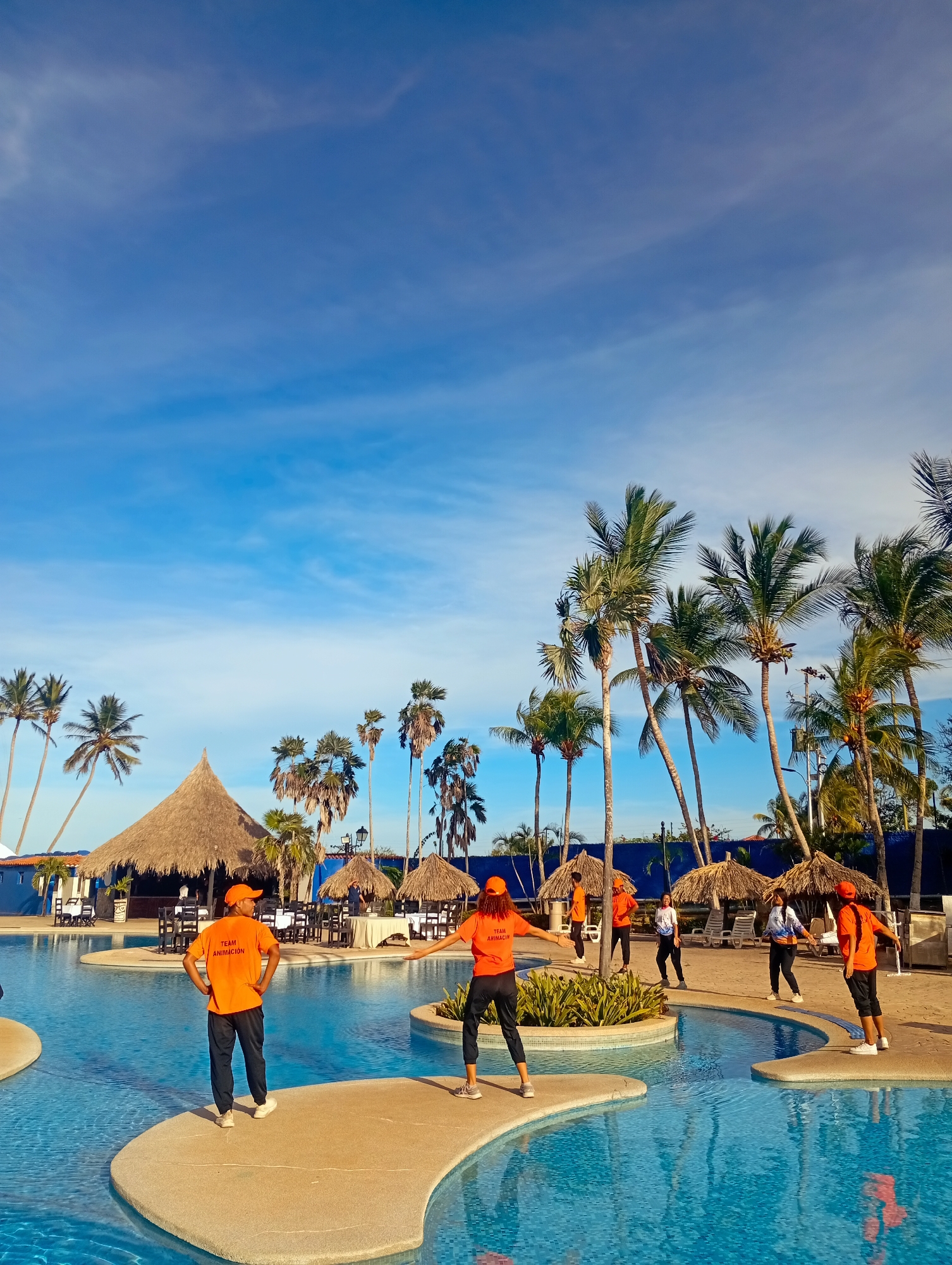 The scene is set at a resort in Playa El Agua, Venezuela, featuring a large swimming pool with several small islands connected by walkways. A group of people wearing orange shirts and black pants are gathered on the islands, seemingly leading or participating in some form of exercise or activity. The shirts have the words "TEAM ANIMACIÓN" printed on them.
The environment includes palm trees scattered around the pool area, thatched-roof cabanas, and a blue building structure. The sky is bright blue with scattered clouds, suggesting a sunny day. Tables and chairs are visible under one of the cabanas, indicating a dining or relaxation area. The pool water is clear and turquoise.