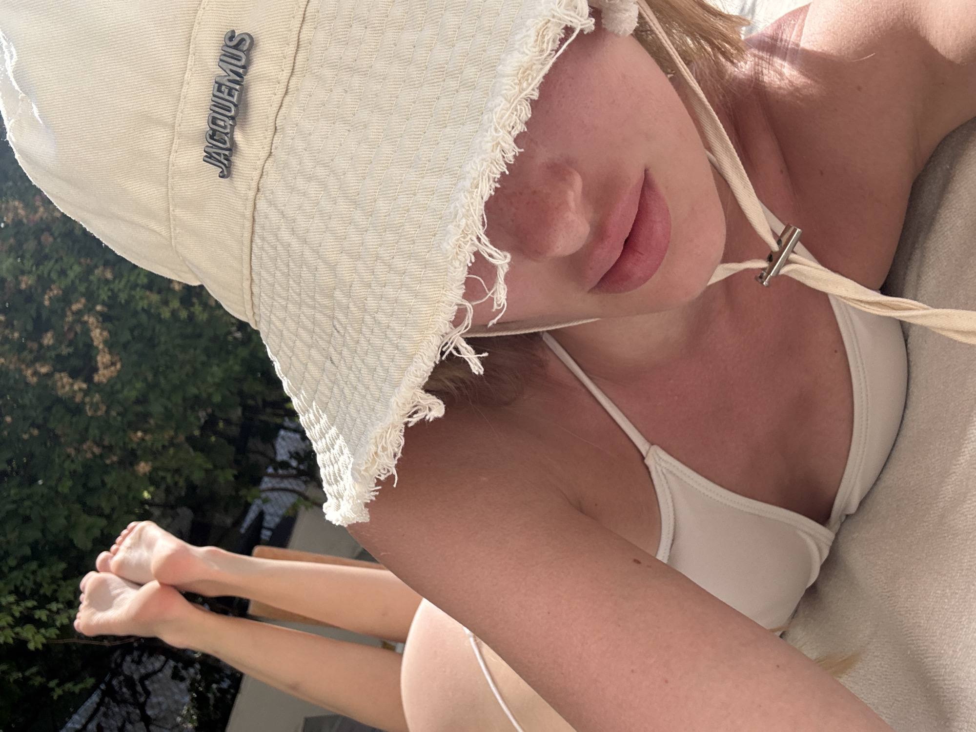 A young Caucasian woman with blonde hair is lying on a lounge chair outdoors, with her legs raised. She is wearing a white bikini and a cream-colored bucket hat with the "Jacquemus" logo. The hat is partially obscuring her face, but her lips and nose are visible. Her feet are in focus in the upper left corner of the image. The background shows green foliage, suggesting a natural or garden setting, consistent with a location in Kemer, Turkey, a resort town known for its natural beauty and outdoor leisure activities. The lighting indicates it is daytime, likely sunny.
